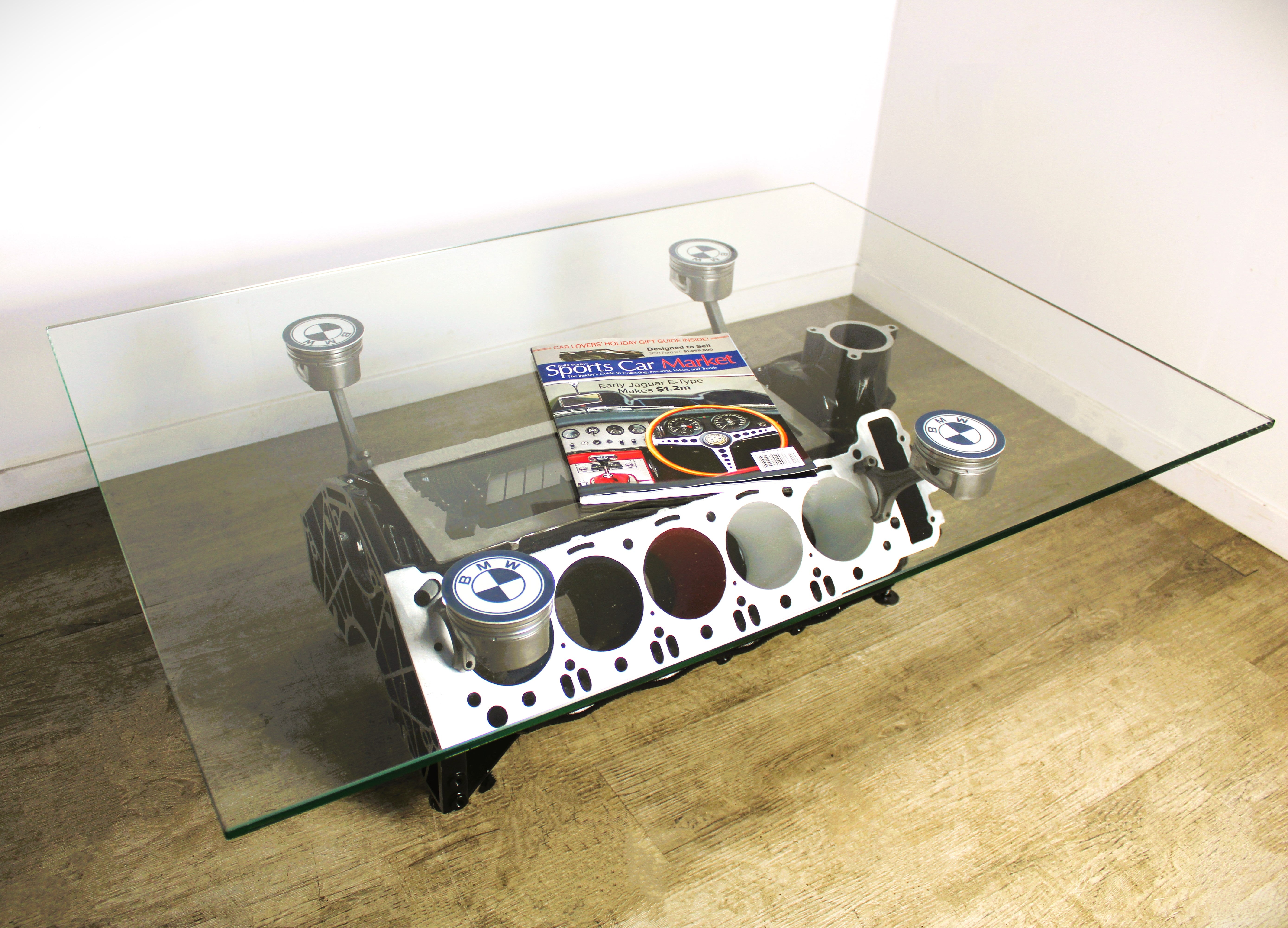 BMW M Series V10 engine block coffee table painted in the BMW M-Power color scheme with a rectangular glass top, the M-Power logo displayed across the piece, and the BMW logo on each of the four car pistons. A sports car magazine rests on top.