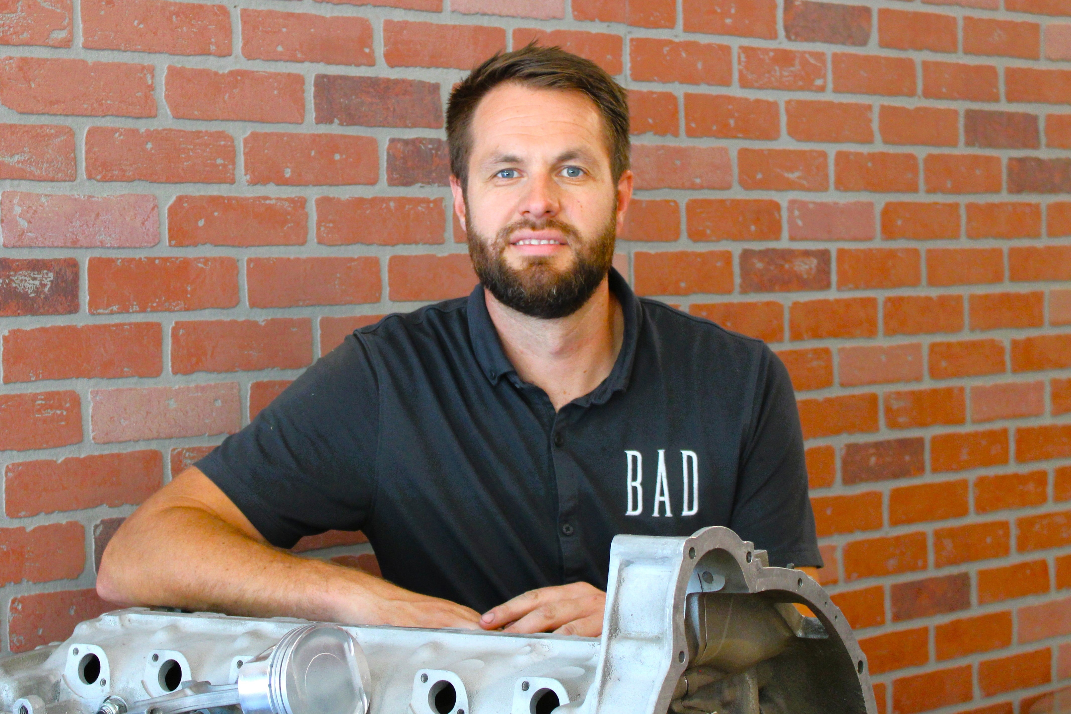 Man wearing a black shirt with 'BAD' text, standing against a brick wall with mechanical components.