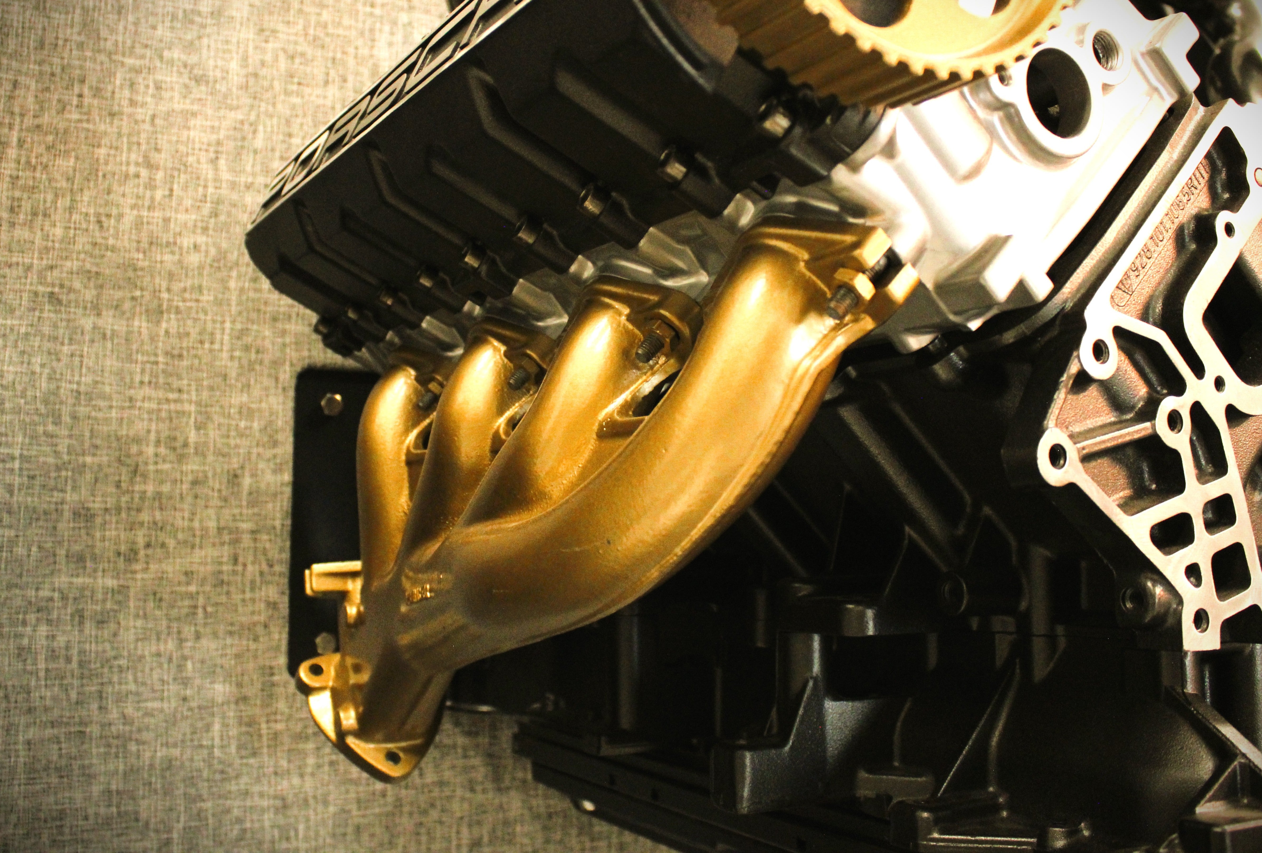 A close-up of a metallic gold exhaust manifold attached to a black and silver engine block, photographed against a textured gray fabric background.