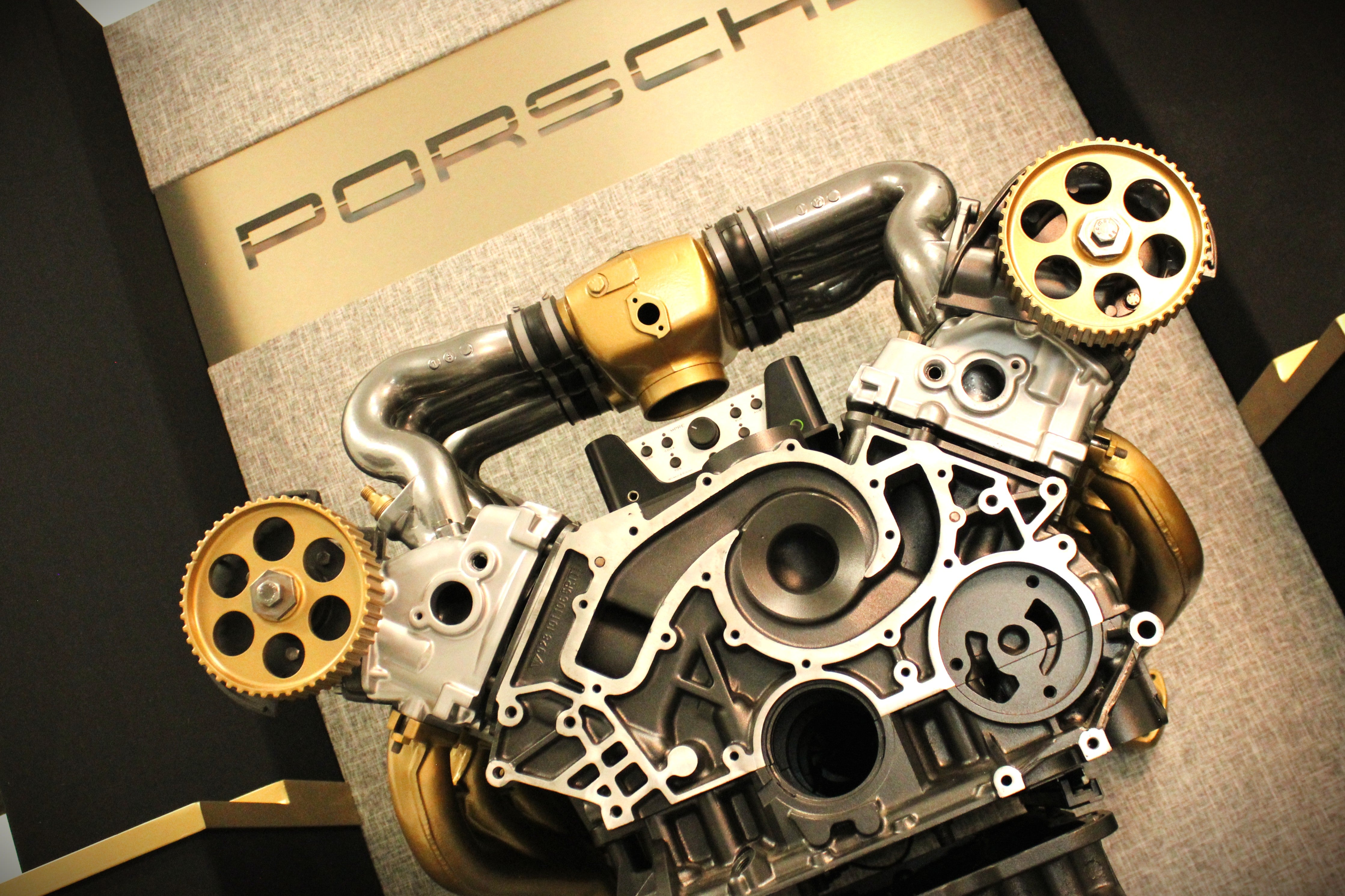 A close-up of a Porsche engine display mounted on a branded display stand with the Porsche logo visible in the background.