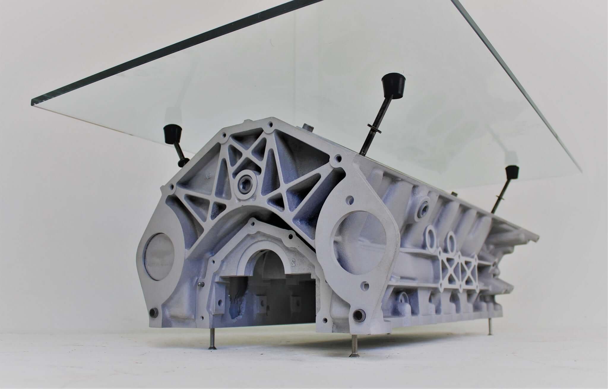 Coffee tables handcrafted from genuine car engines with various colored finishes, logos displayed, and glass top shapes.