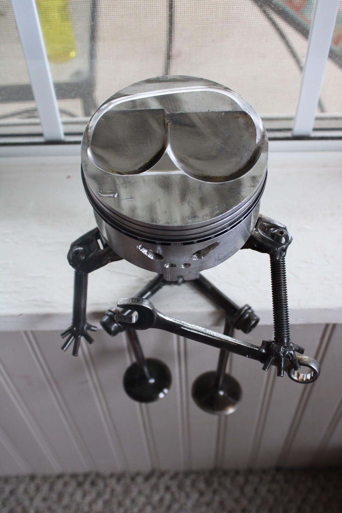 Handmade figurine man made entirely out of a car engine piston and other automotive components, sitting down, polished, and with a glossy finish holding various car parts.