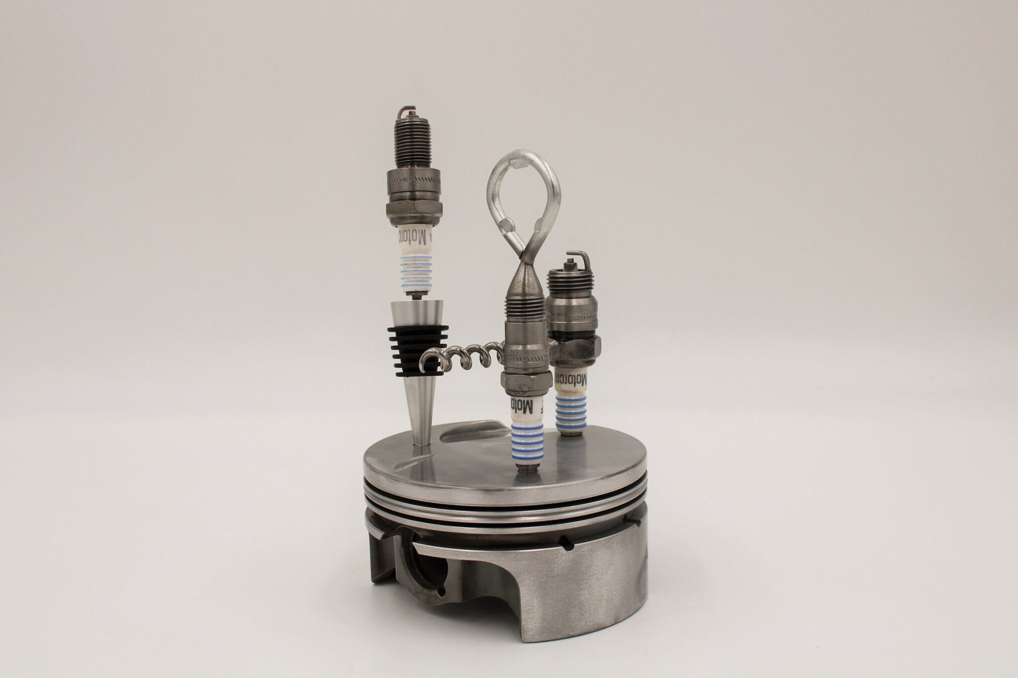 Barware set handcrafted from genuine car engine components - a polished car engine piston holder with a bottle opener, corkscrew, and stopper made from spark plugs.