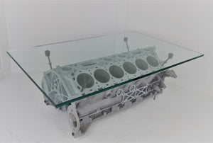 Engine block coffee table painted light gray, with its rectangular glass top being held up by car engine pistons.