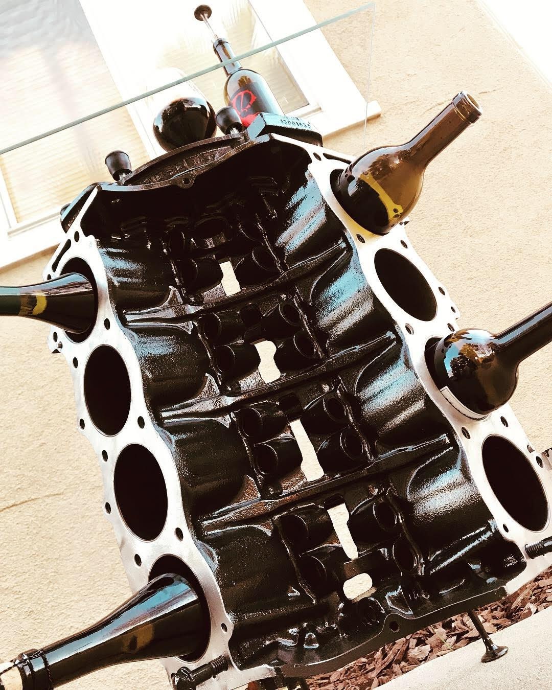 Lower view of an engine block wine rack finished in black with a square glass top, wine bottles stored in its side and a wine bottle and glass on top.