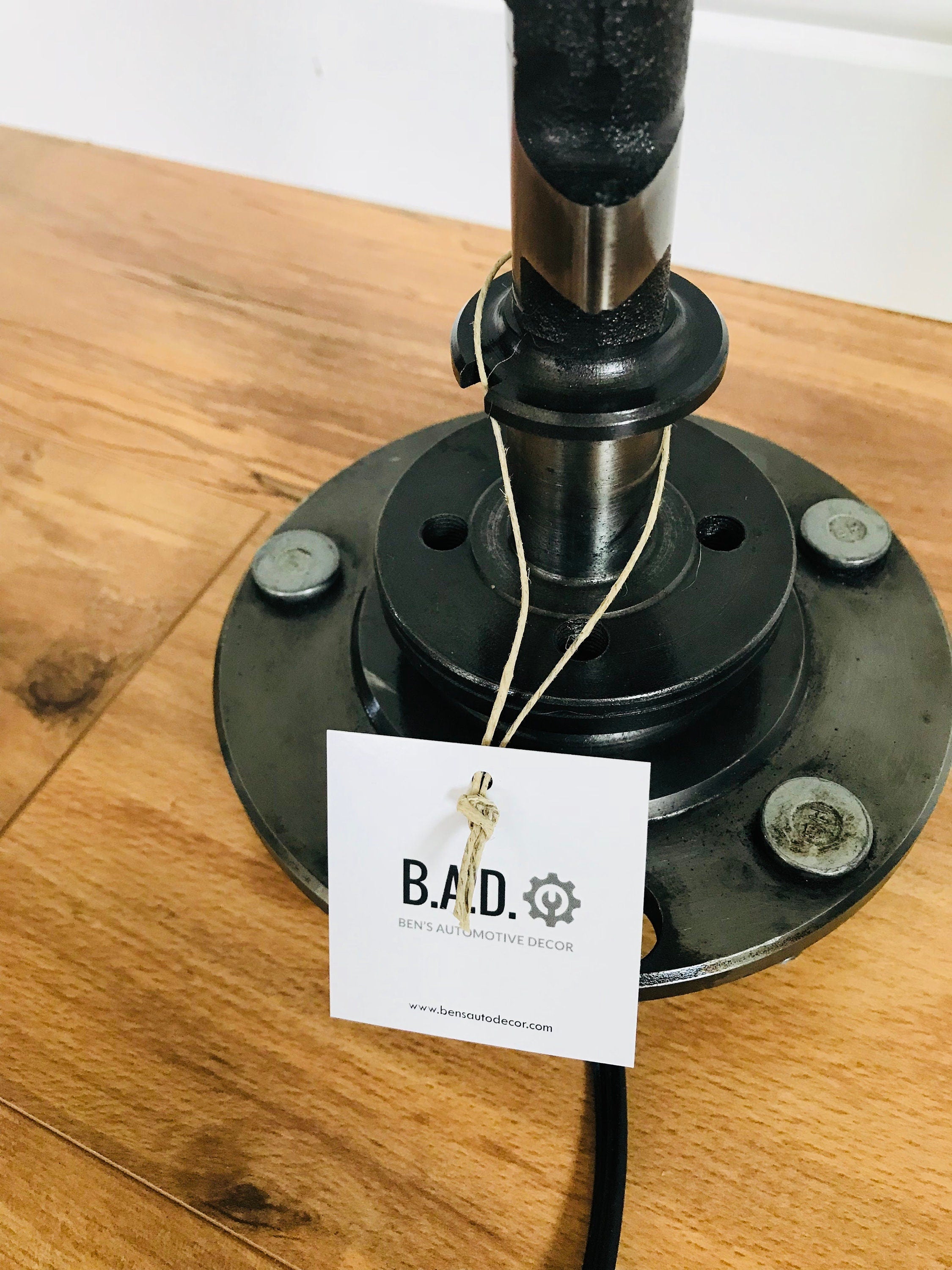 The base of a silver lamp made out of a car's camshaft with a tag reading, "B.A.D., Ben's Automotive Decor".