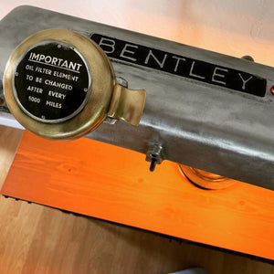 Close-up view of a desk lamp made from a car's valve cover, the Bentley logo displayed on top and including its oil filter holder, reading, "Important - Oil filter element to be changed after every 5000 miles"