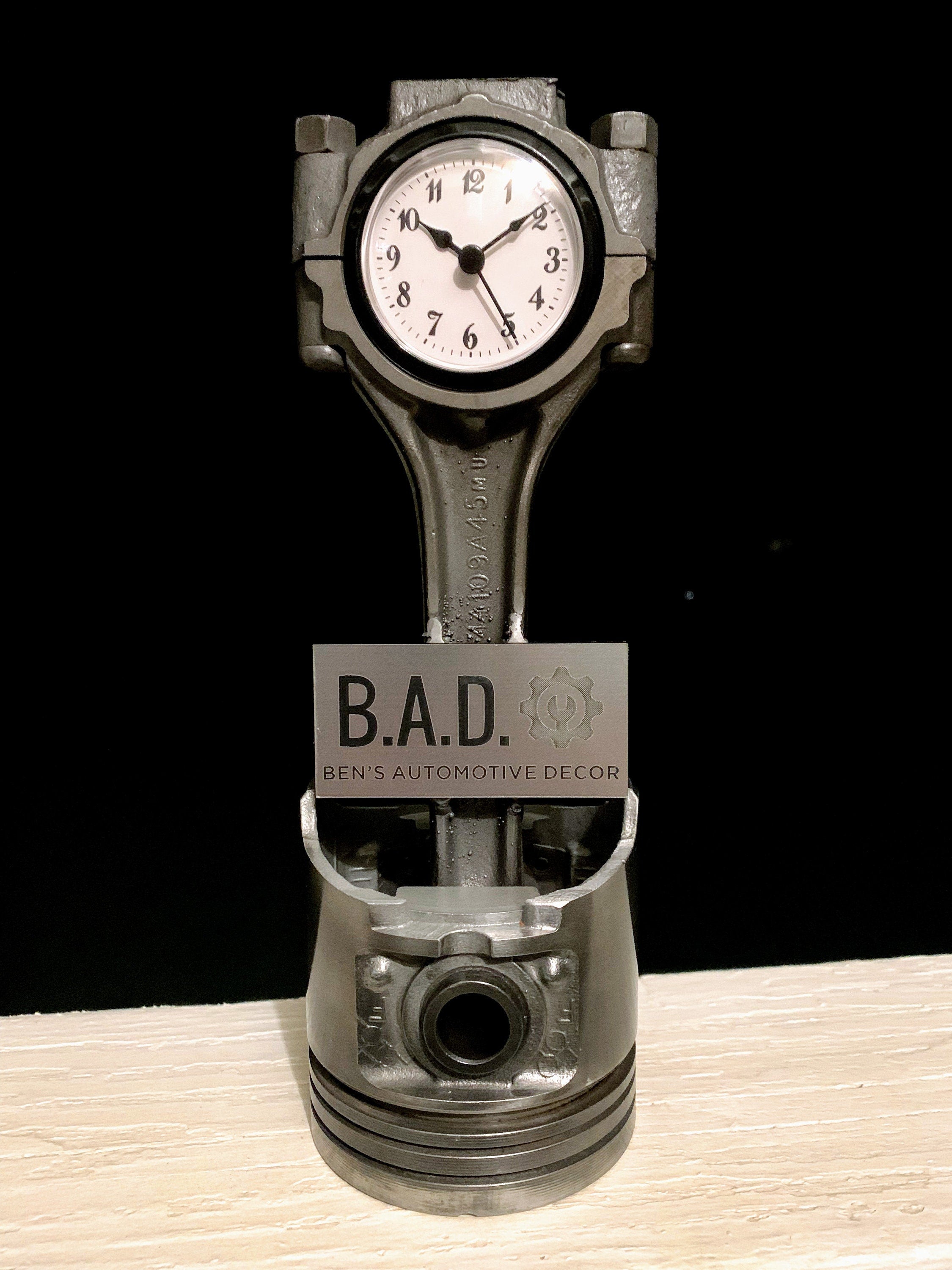 Clock made out of a car engine piston with a custom plaque, reading "B.A.D., Ben's Automotive Decor".