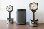 Load image into Gallery viewer, Two car piston clocks beside a speaker on a table. Left clock is finished in gunmetal gray with a silver clock ring, and the right clock has a patina finish with a gold clock ring.
