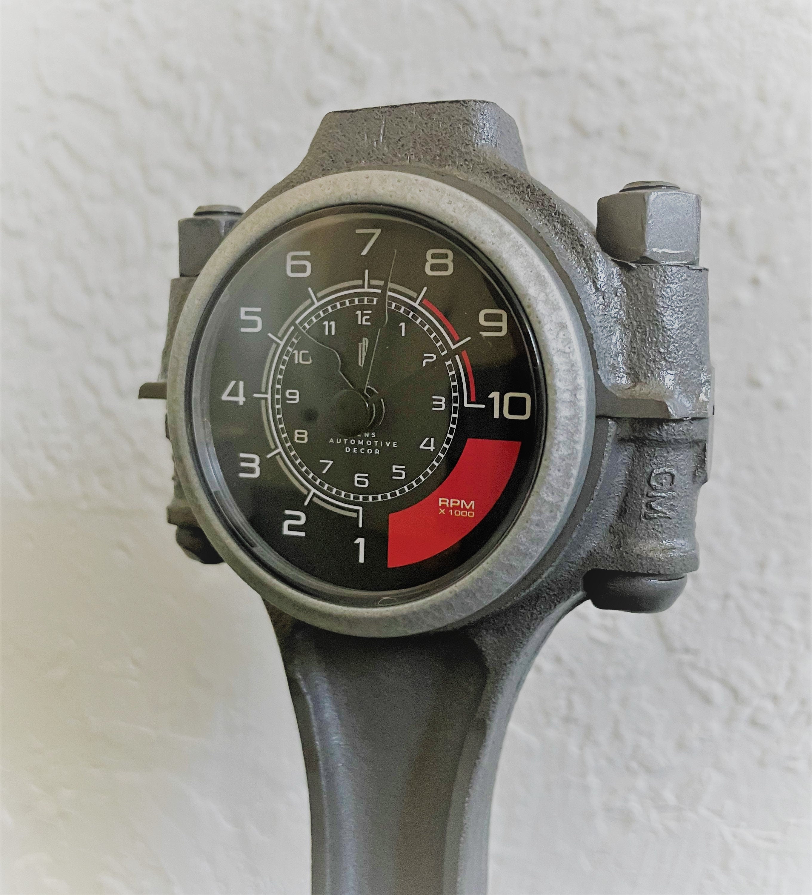 Close-up view of a car piston clock finished in gunmetal gray with a silver clock ring and black and red RPM clock face.