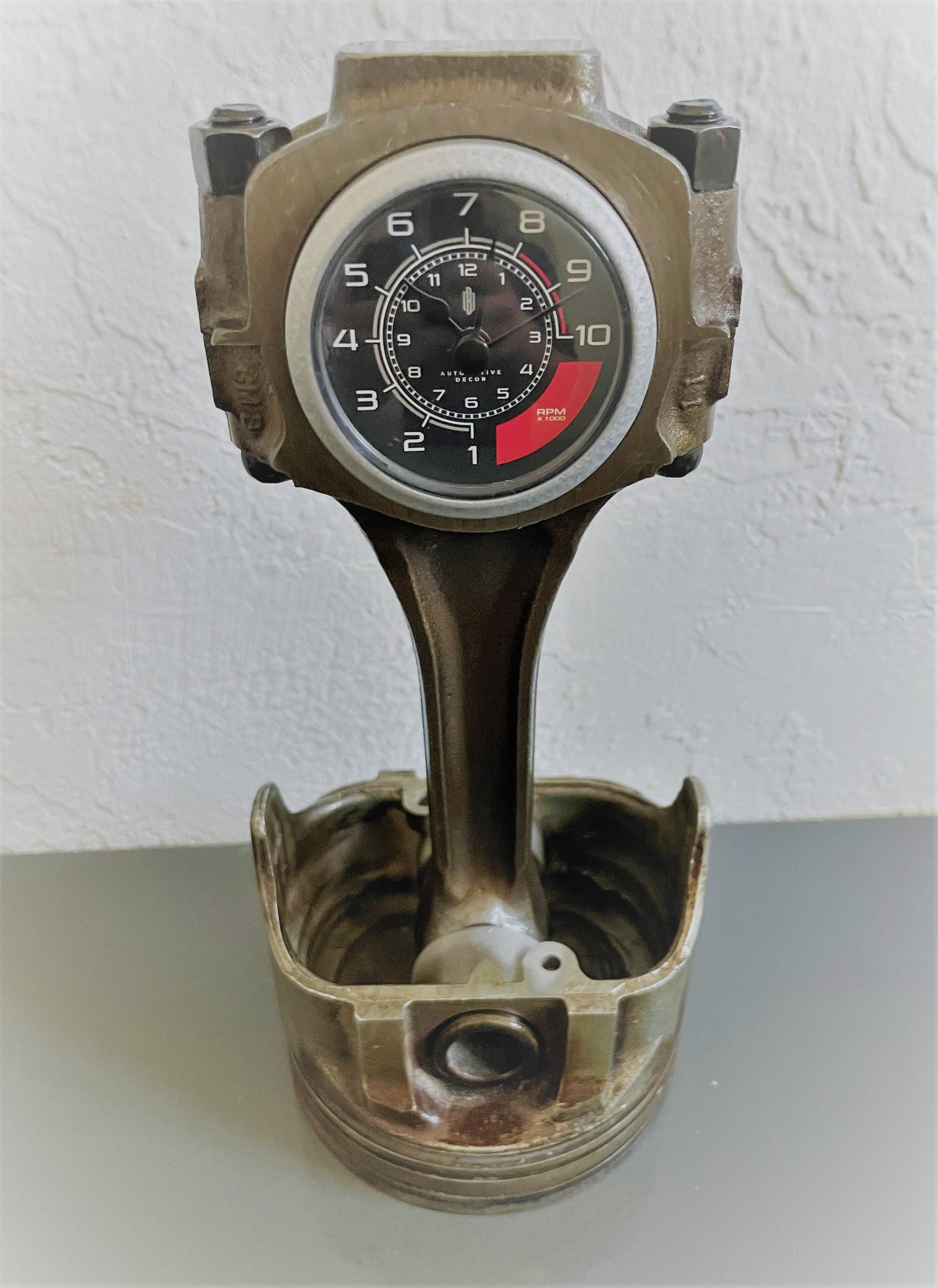 A car piston clock in a patina finish with a silver clock ring and black and red RPM clock face.