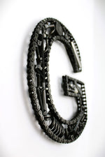Load image into Gallery viewer, A letter G made out of real car parts, outlined with a timing chain.
