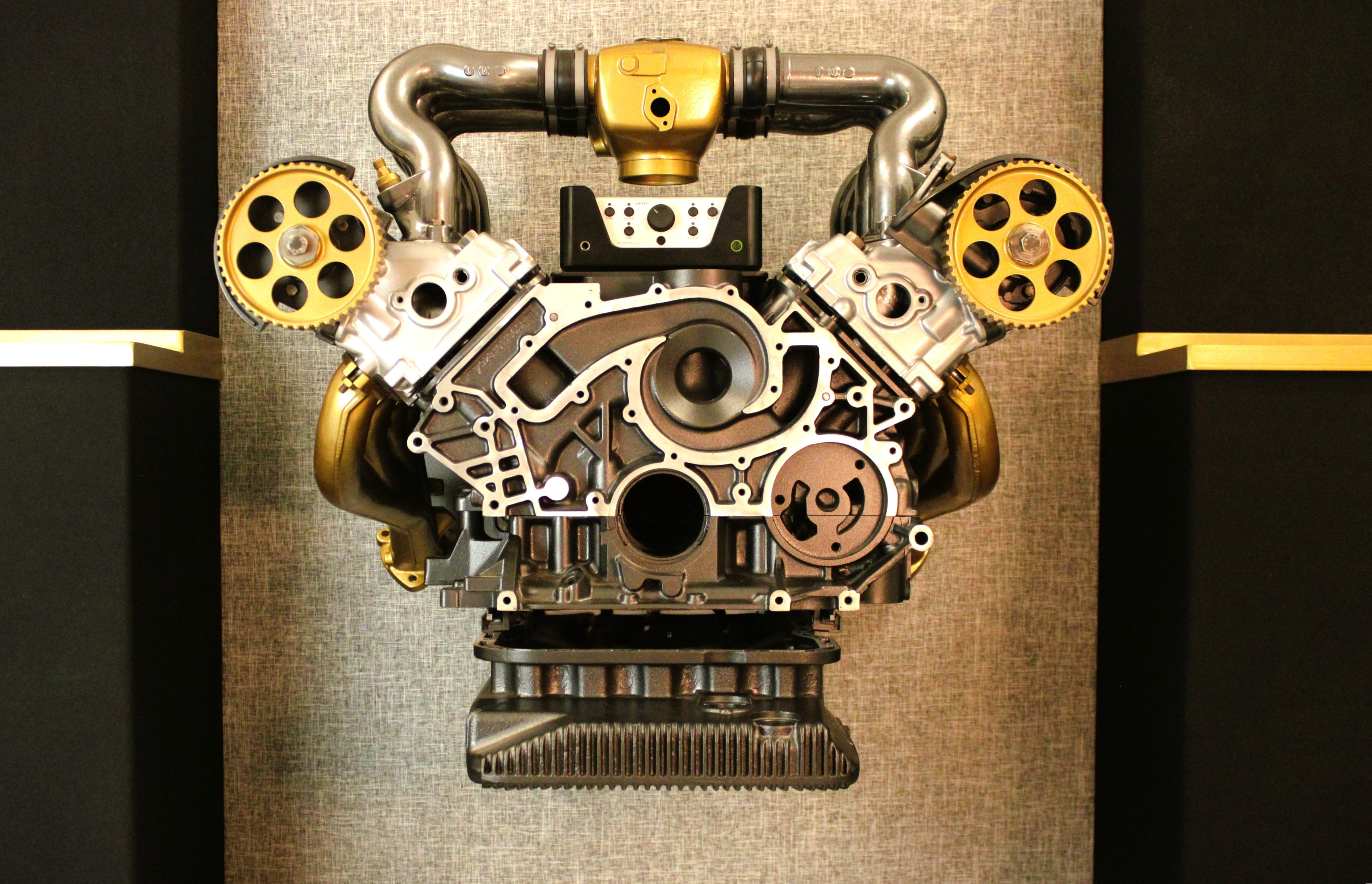 A detailed, cut-open V8 engine display mounted on a textured beige wall, flanked by dark display cabinets. The engine features prominent gold-colored camshaft pulleys, polished silver metal components, and a black crankcase.