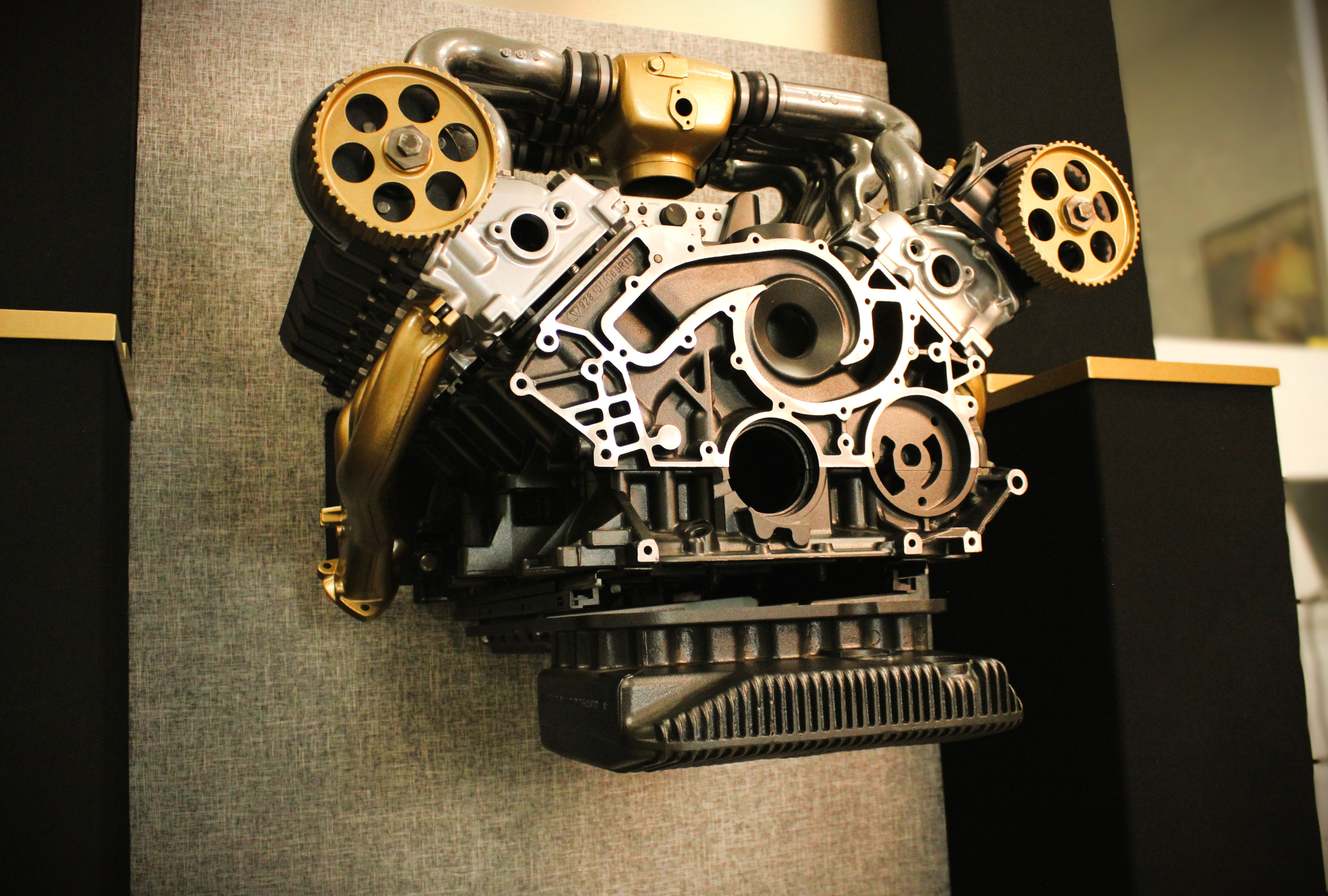 A detailed cross-section of a V8 engine block with gold-colored timing belts and pulleys, mounted on a gray textured display board.