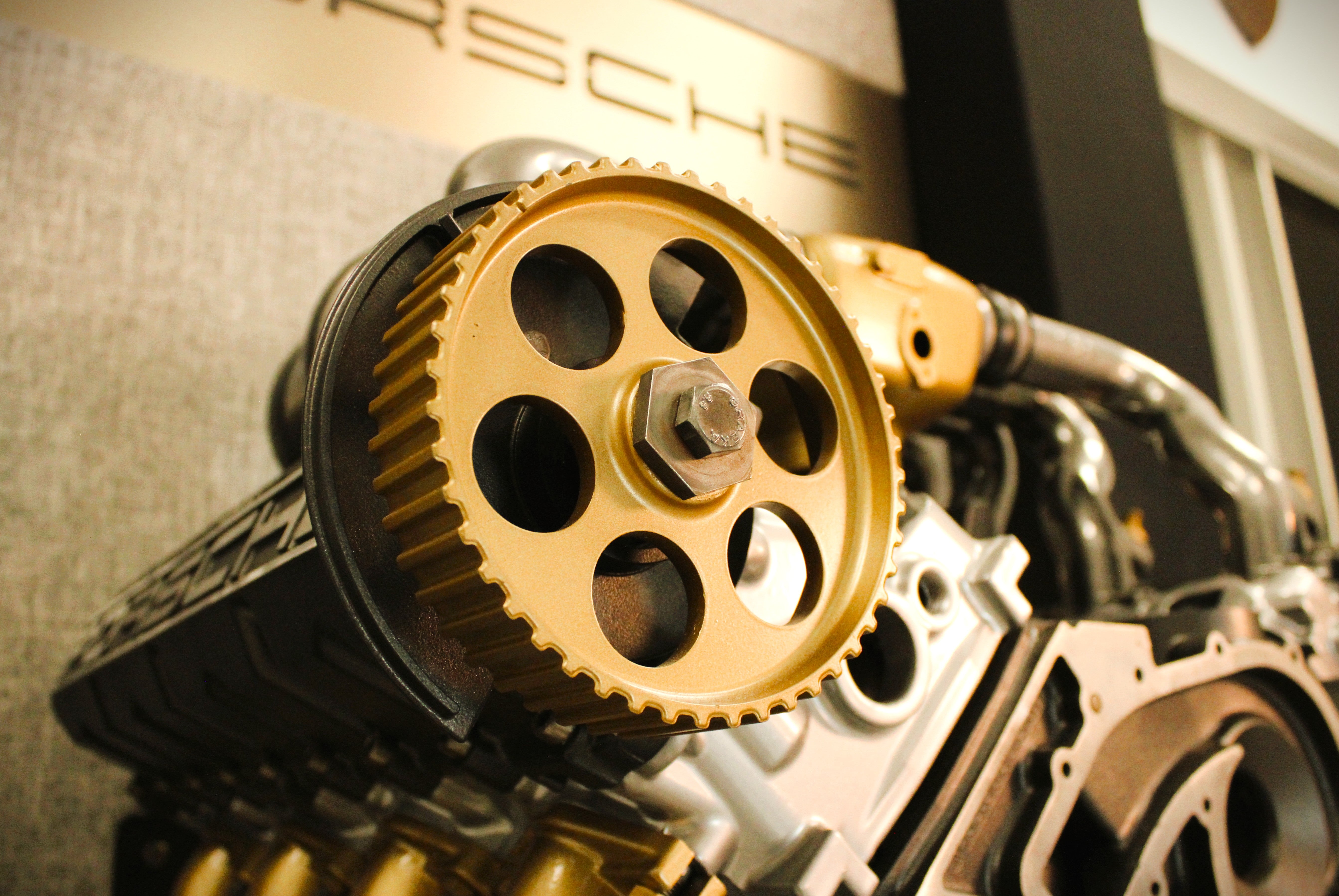 Close-up of a gold and black Porsche engine component, showing a large toothed pulley wheel with a central bolt and surrounding engine parts.