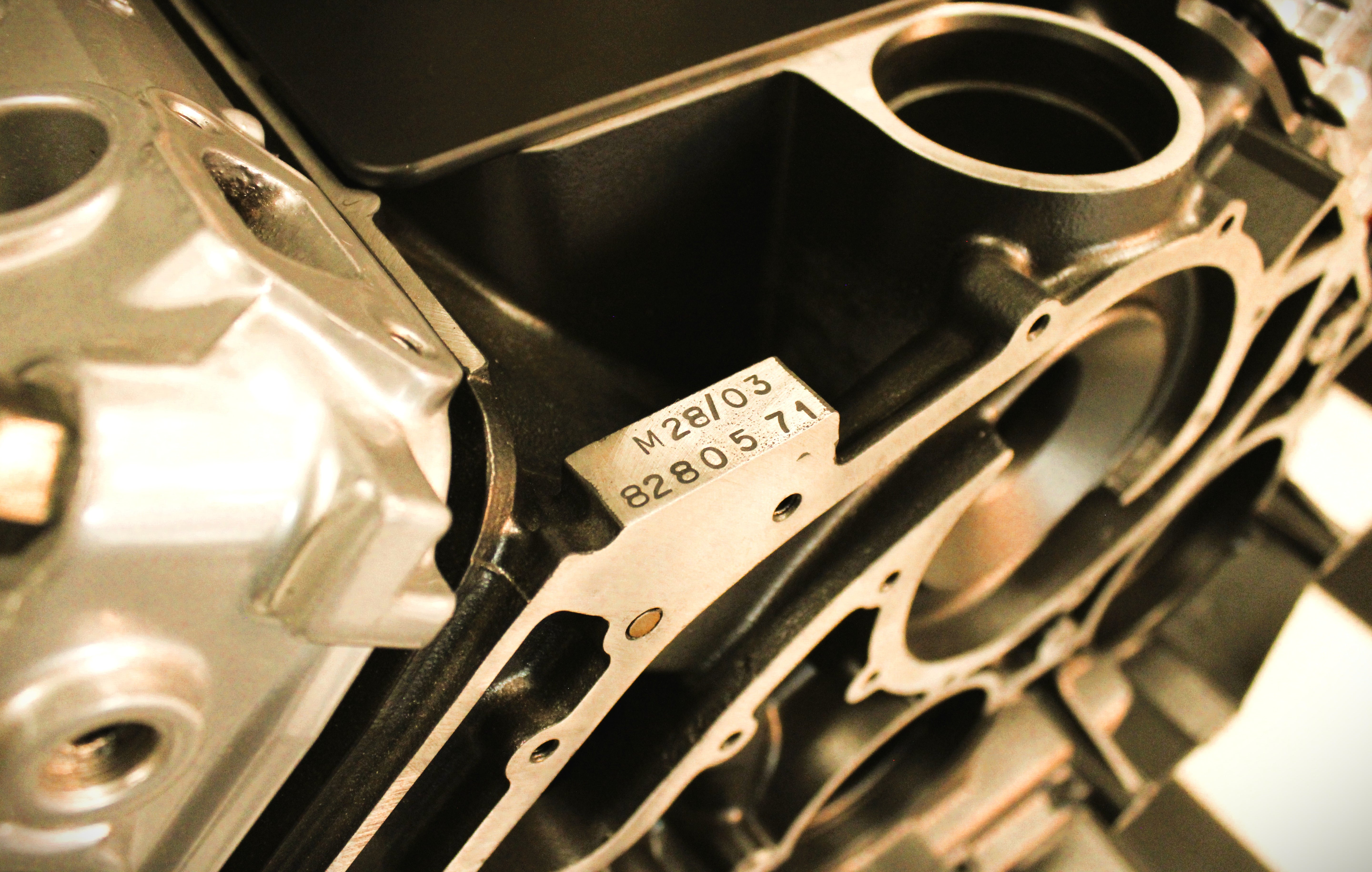 Close-up of a metallic engine block or mechanical housing with a small white sticker showing the code M28J03 82805.71, photographed in warm golden lighting.