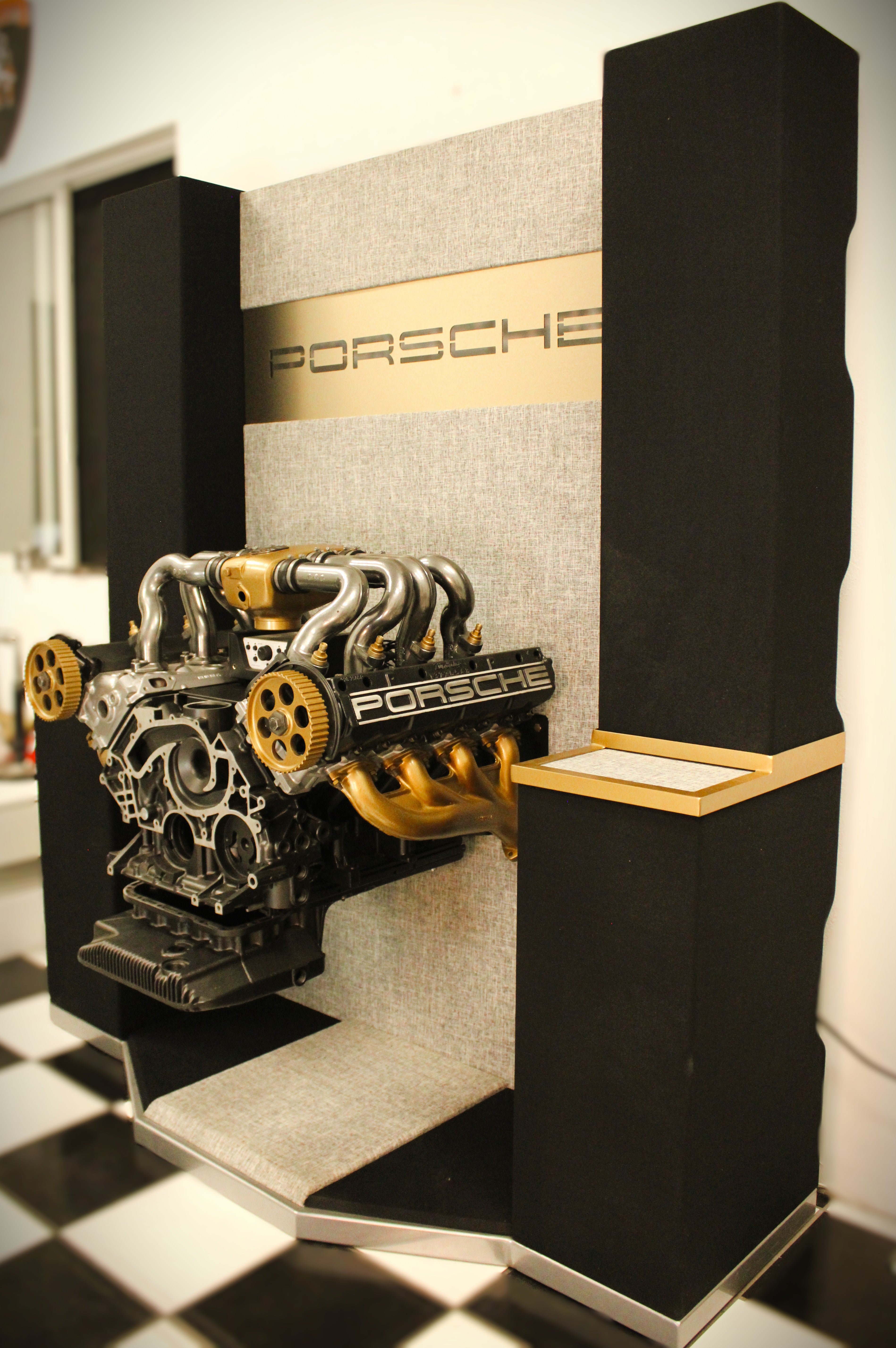 A detailed Porsche engine model mounted on a audio system with a gray fabric background, gold accent strip, and black side panels. The Porsche logo is visible on the engine and the display panel.