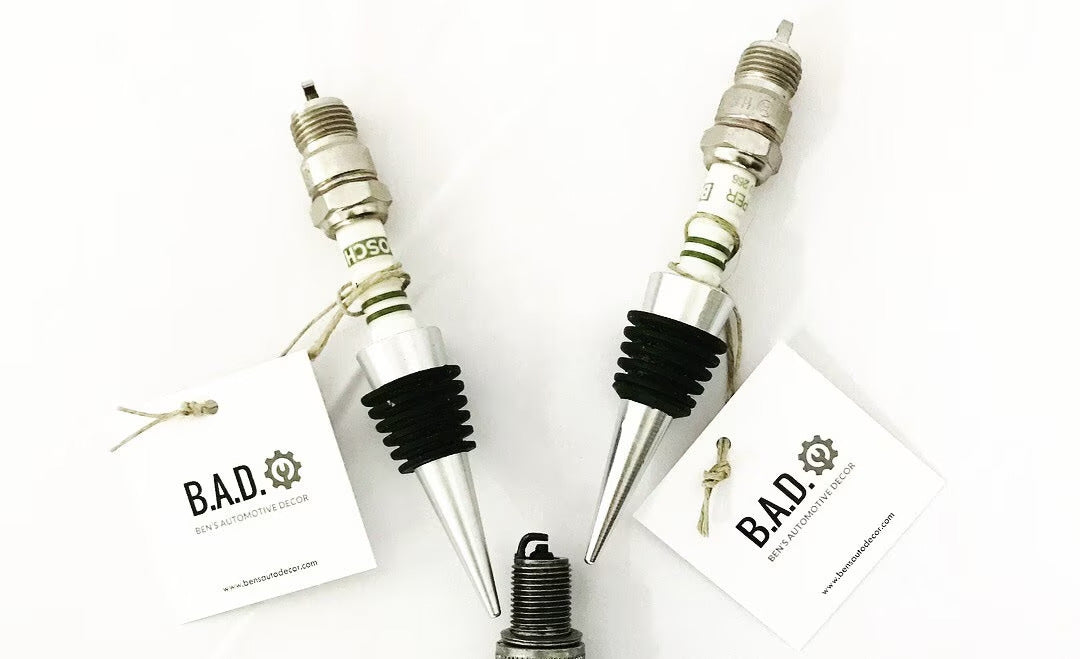 Two bottle stoppers made from car engine spark plugs with tags reading, "B.A.D., Ben's Automotive Decor"