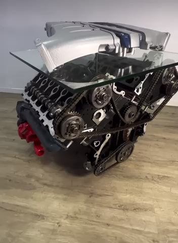 Video of a multicolored Twin Turbo rotating engine block coffee table with several moving parts on wheels, finished with a rectangular glass top.