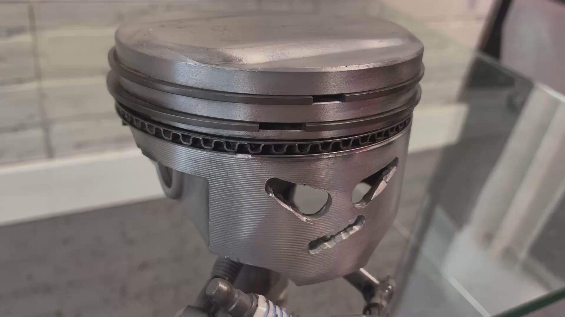 Video of a sitting piston man car part sculpture holding a spark plug.