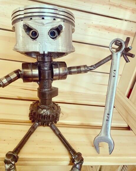 Handmade figurine man made entirely out of a car engine piston and other automotive components, sitting down, polished, and with a glossy finish holding various car parts.