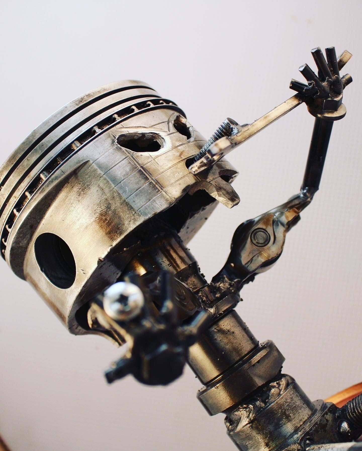 Handmade figurine man made entirely out of a car engine piston and other automotive components, sitting down, polished, and with a glossy finish holding various car parts.