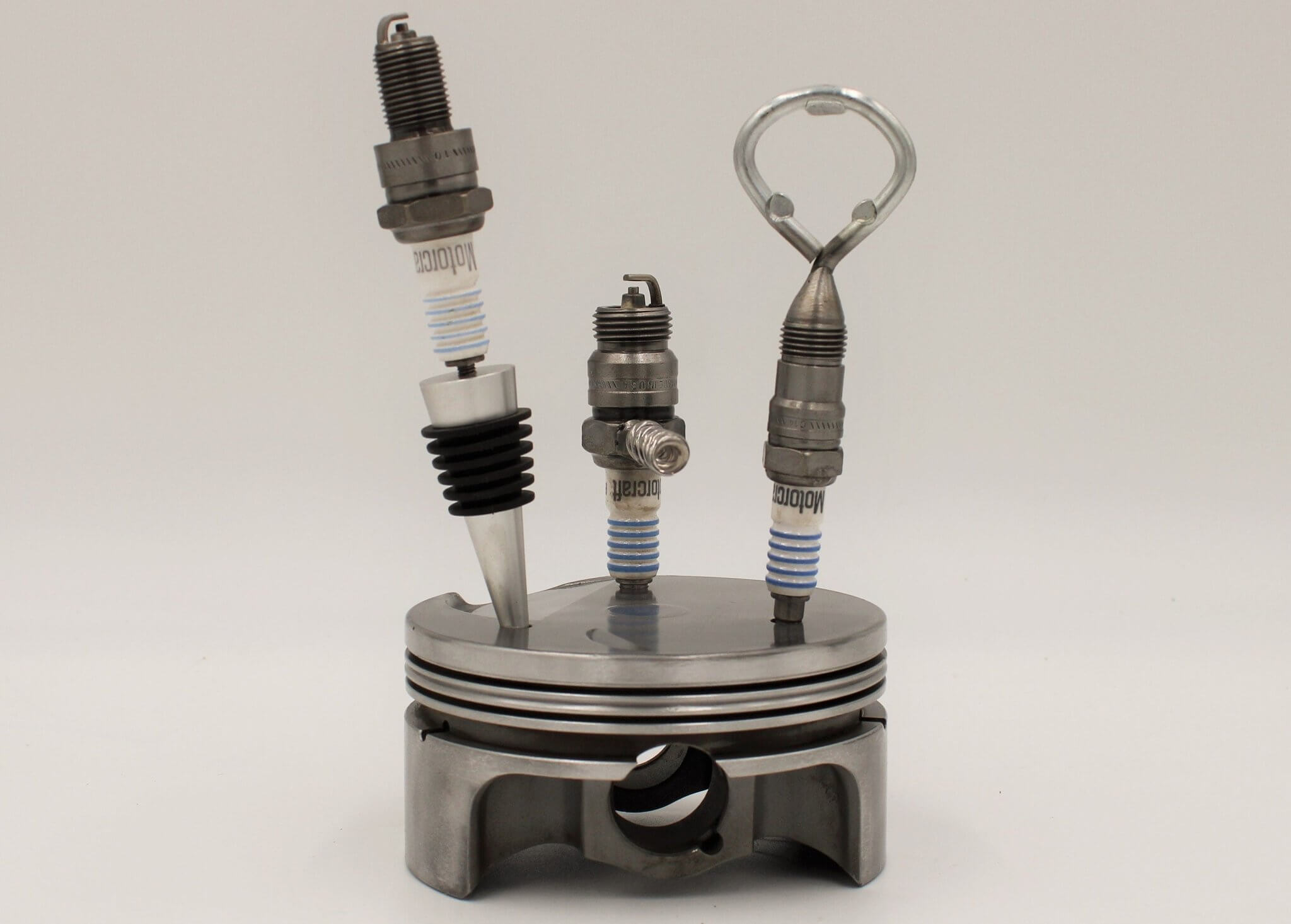 Barware set handcrafted from genuine car engine components - a polished car engine piston holder with a bottle opener, corkscrew, and stopper made from spark plugs.