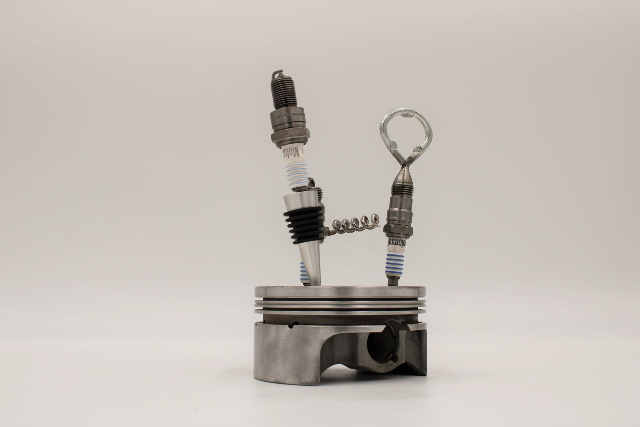 Barware set handcrafted from genuine car engine components - a polished car engine piston holder with a bottle opener, corkscrew, and stopper made from spark plugs.