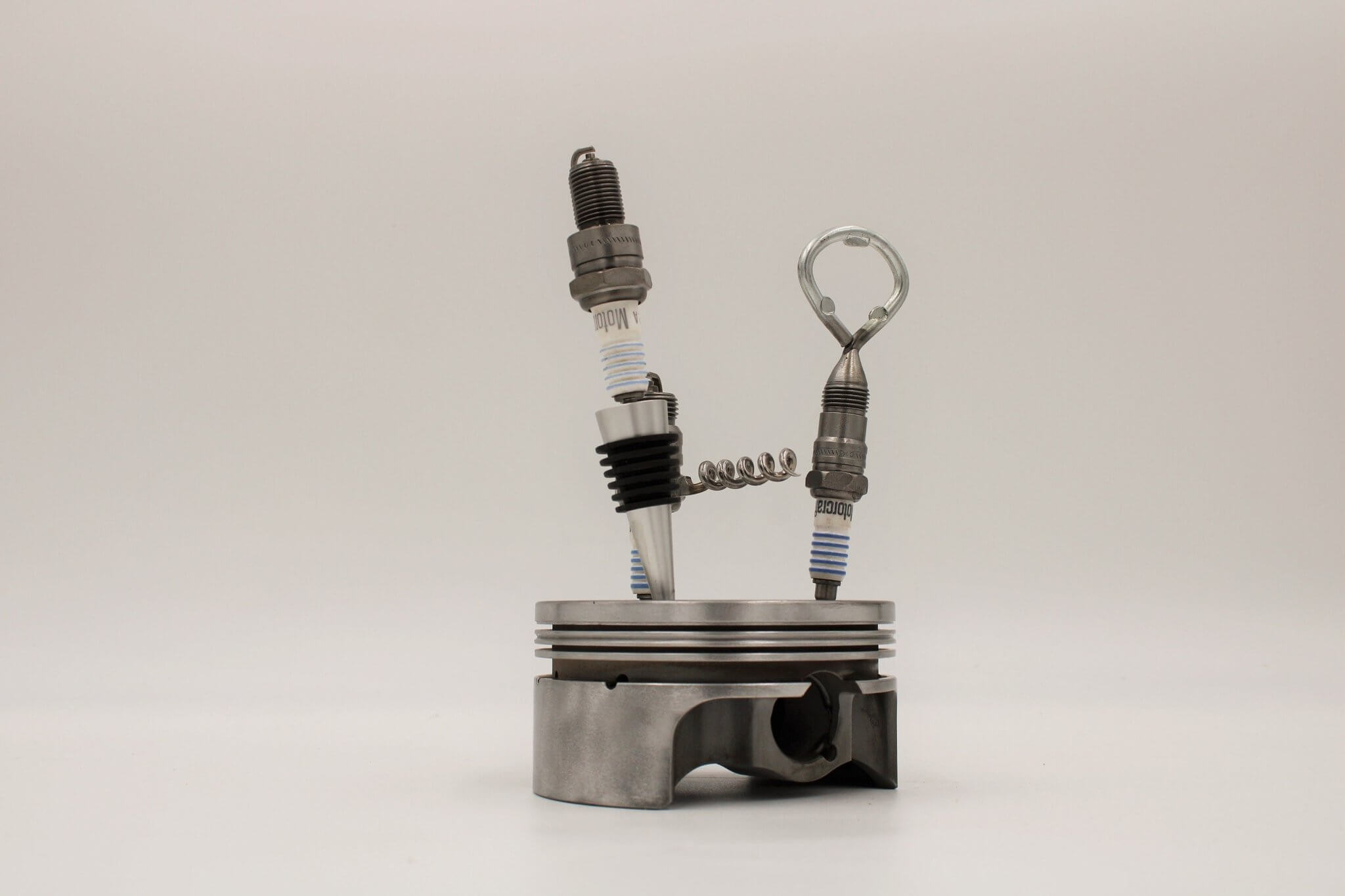 Barware set handcrafted from genuine car engine components - a polished car engine piston holder with a bottle opener, corkscrew, and stopper made from spark plugs.