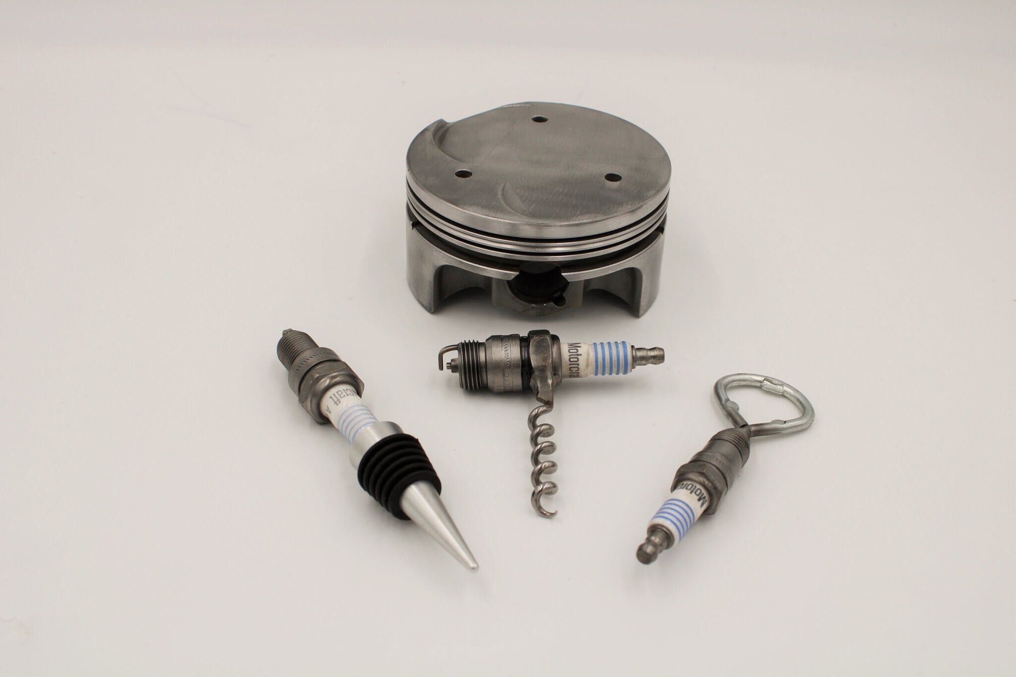 Barware set handcrafted from genuine car engine components - a polished car engine piston holder with a bottle opener, corkscrew, and stopper made from spark plugs.
