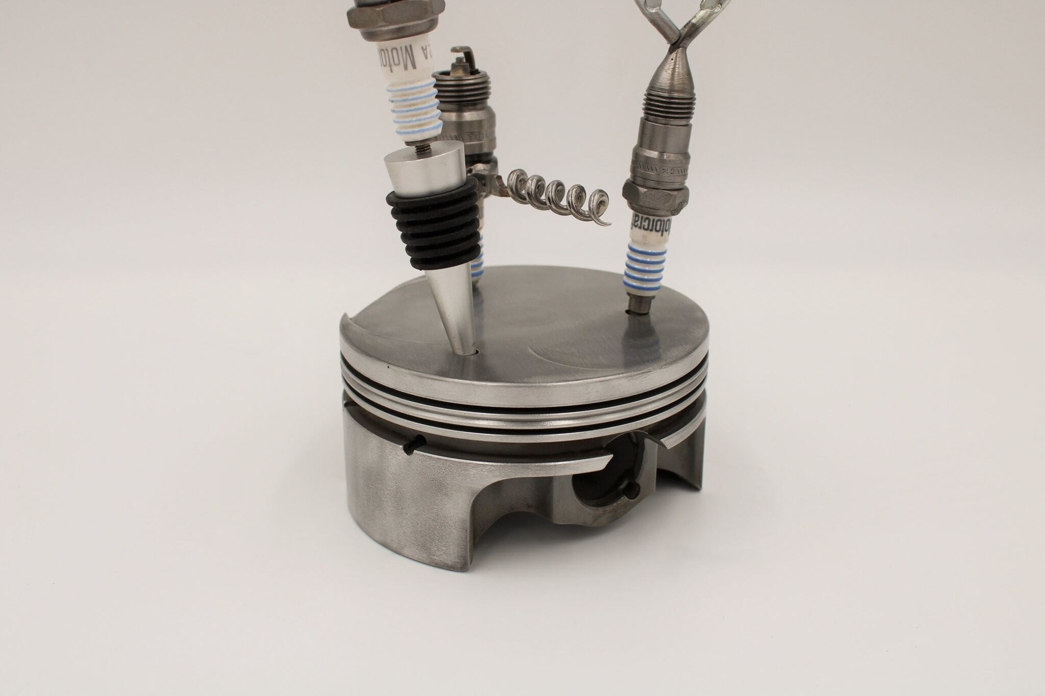 Barware set handcrafted from genuine car engine components - a polished car engine piston holder with a bottle opener, corkscrew, and stopper made from spark plugs.