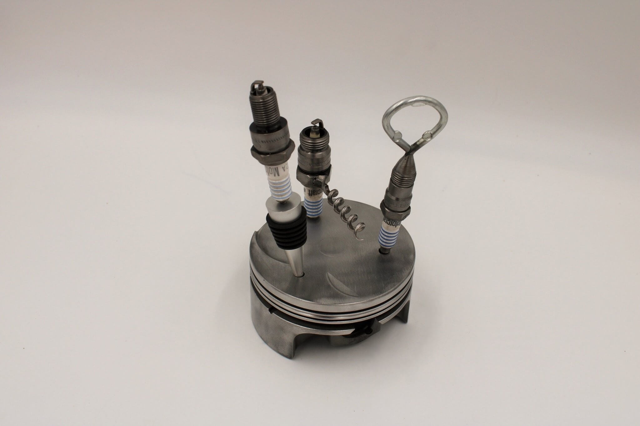 Barware set handcrafted from genuine car engine components - a polished car engine piston holder with a bottle opener, corkscrew, and stopper made from spark plugs.
