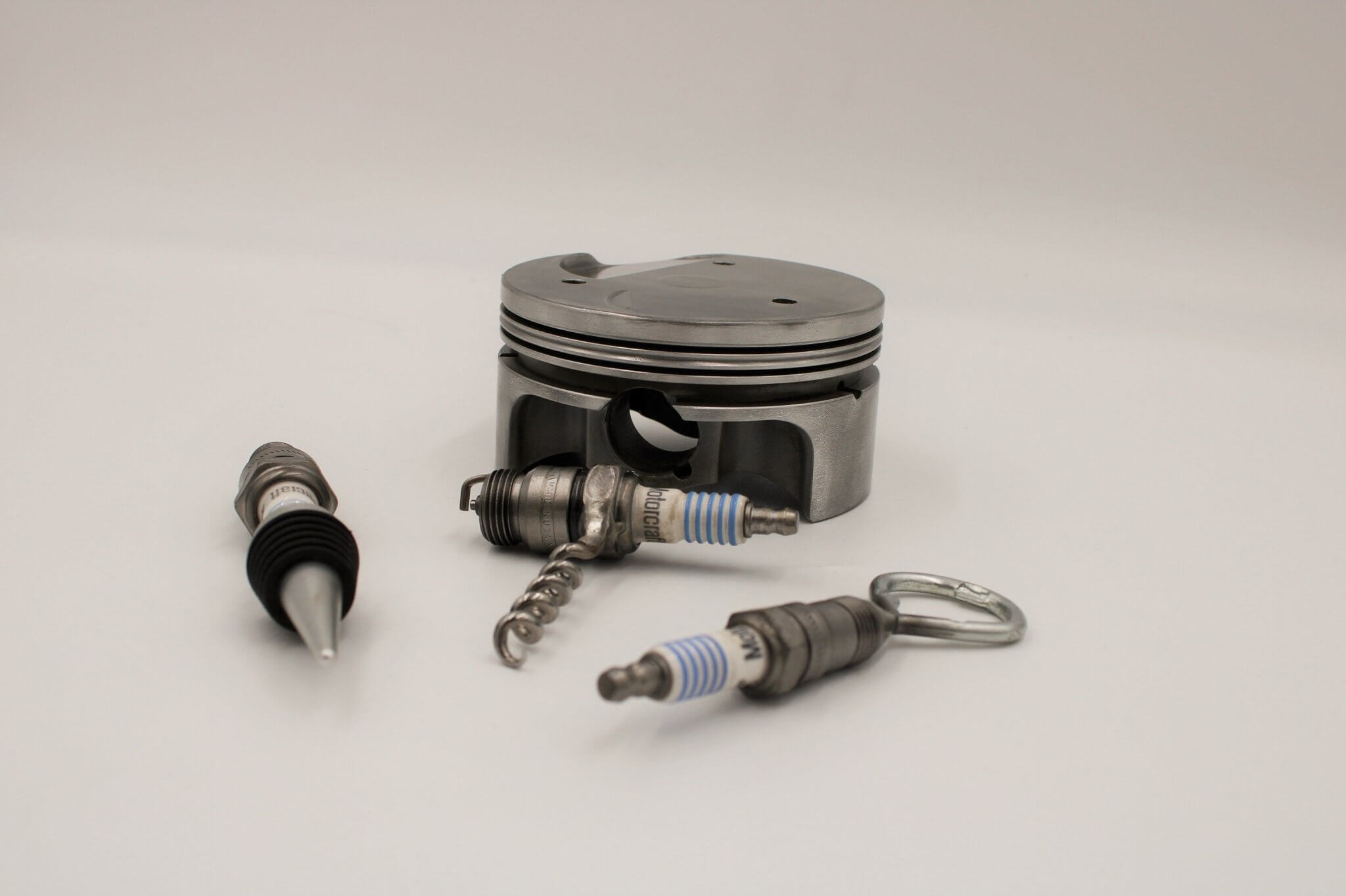 Barware set handcrafted from genuine car engine components - a polished car engine piston holder with a bottle opener, corkscrew, and stopper made from spark plugs.