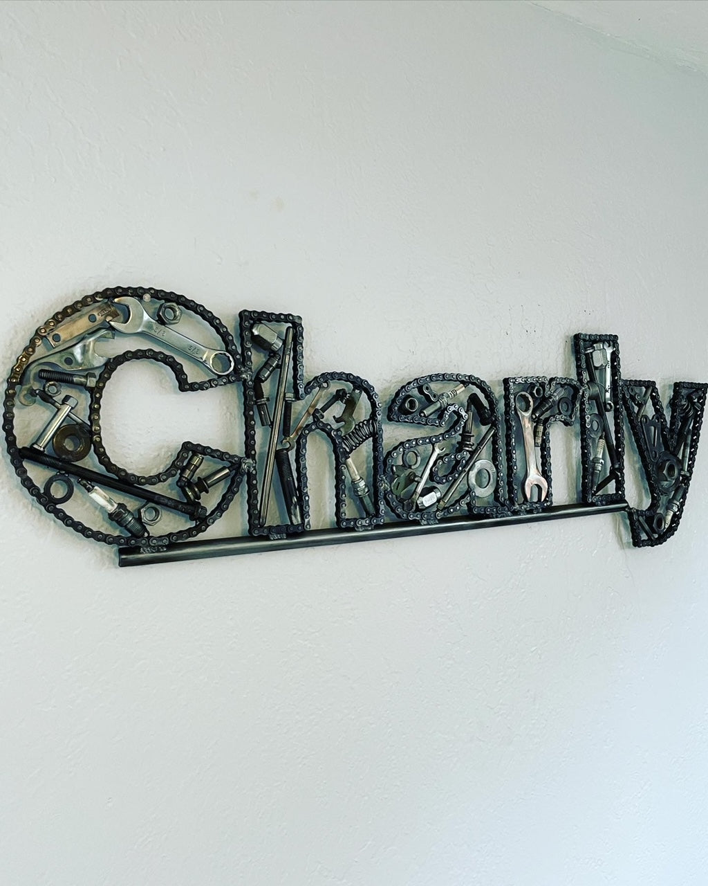 A name sign spelling "Charly" hanging on a wall, each letter made out of car parts and outlined with timing chain.