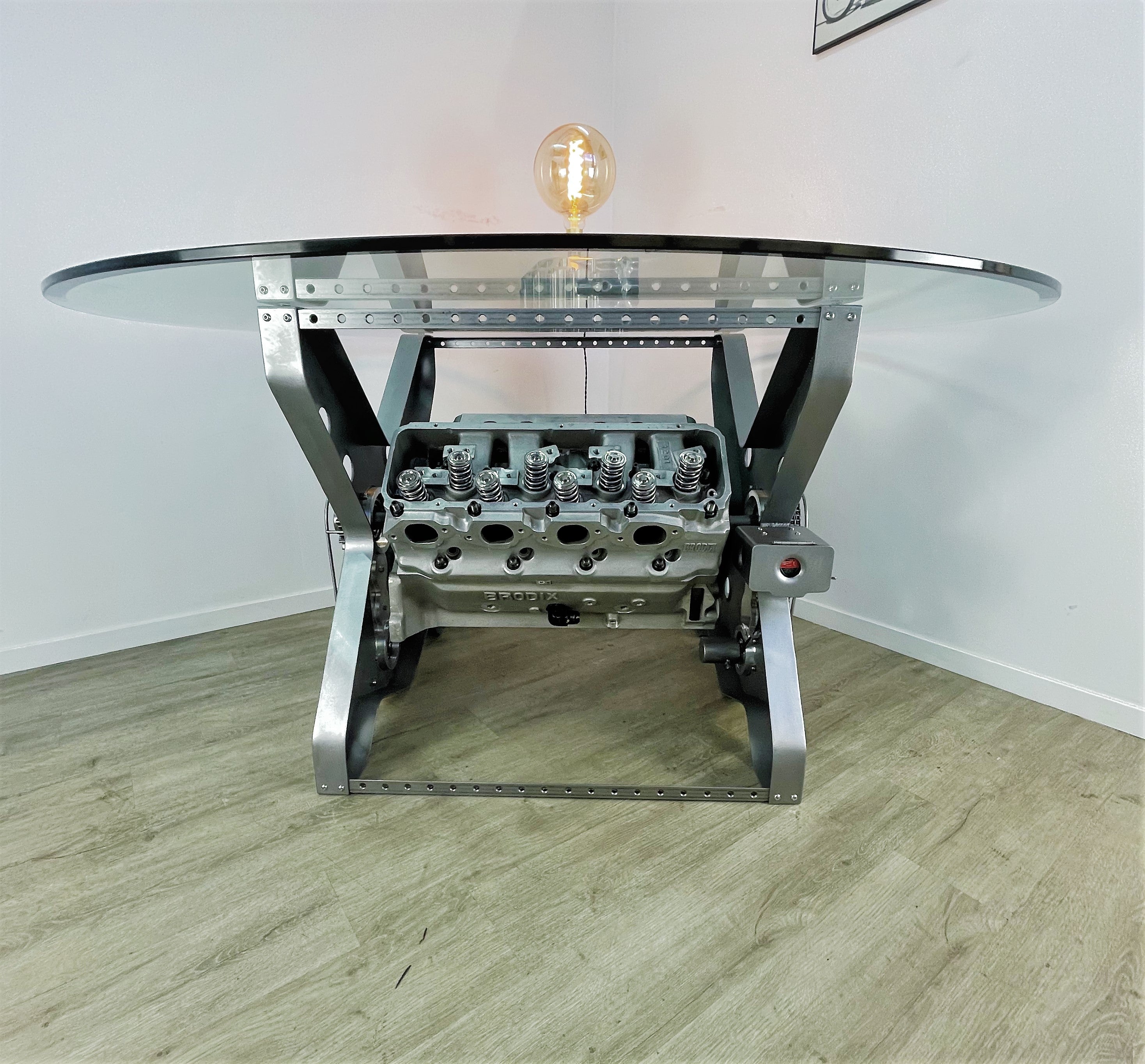 X-frame rotating engine dining table with a round glass top and car part lamp on top.