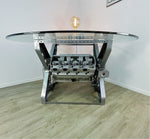 Load image into Gallery viewer, X-frame rotating engine dining table with a round glass top and car part lamp on top.
