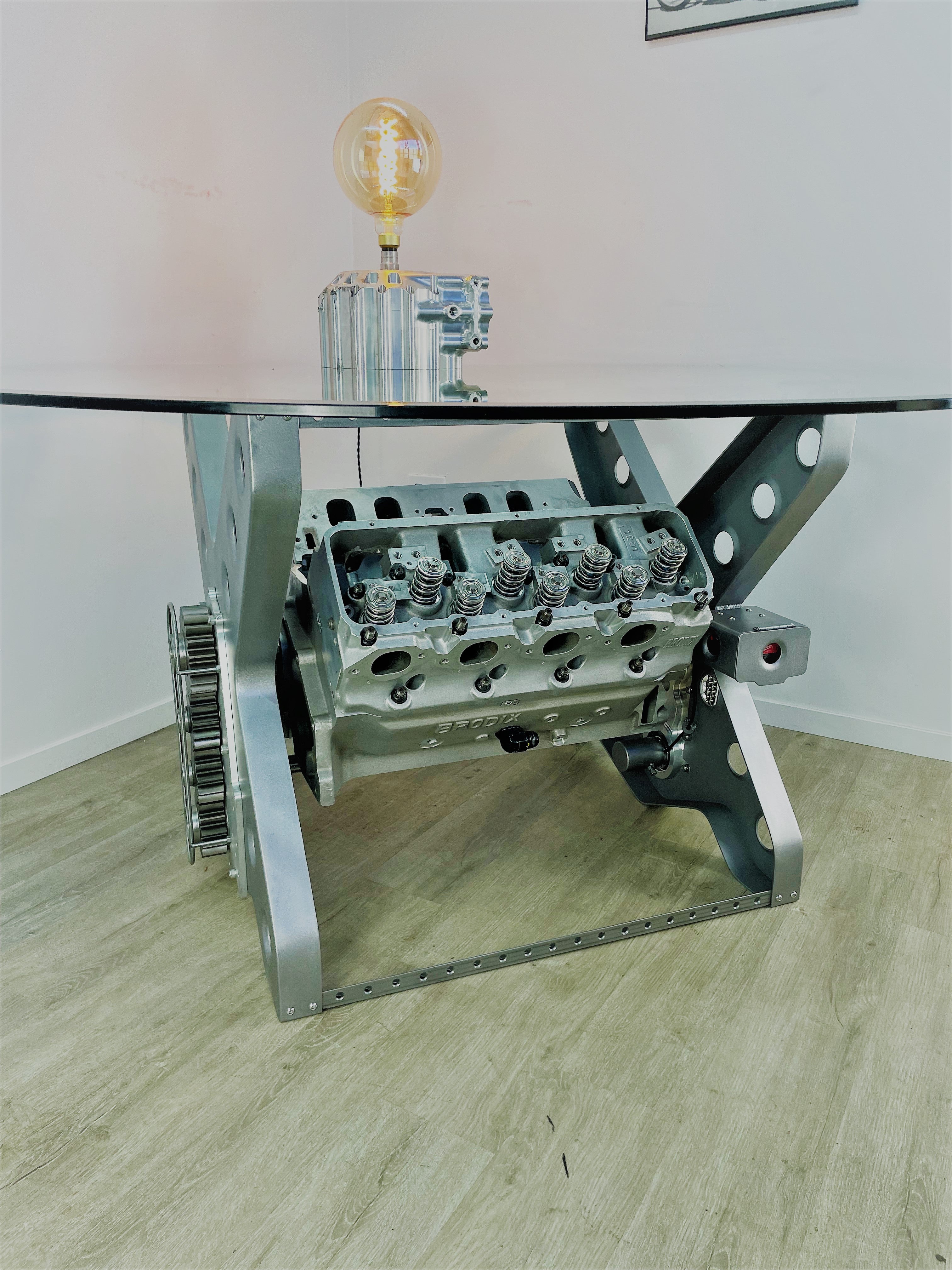 X-frame rotating engine dining table with a round glass top and car part lamp on top.