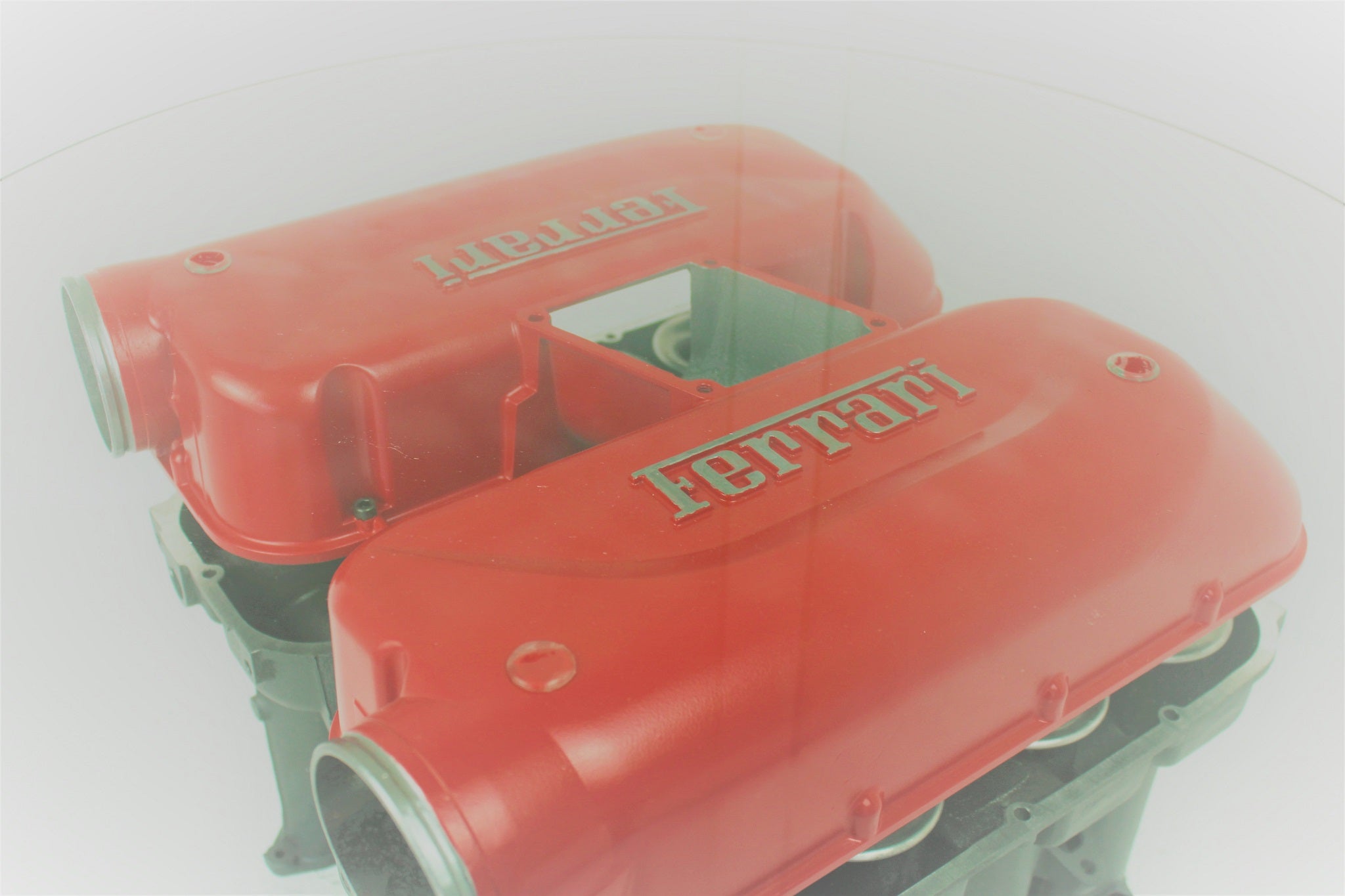 Birds-eye view of a Ferrari intake manifold table, finished in red and gunmetal gray with a round glass top. The Ferrari logo is displayed on both sides.