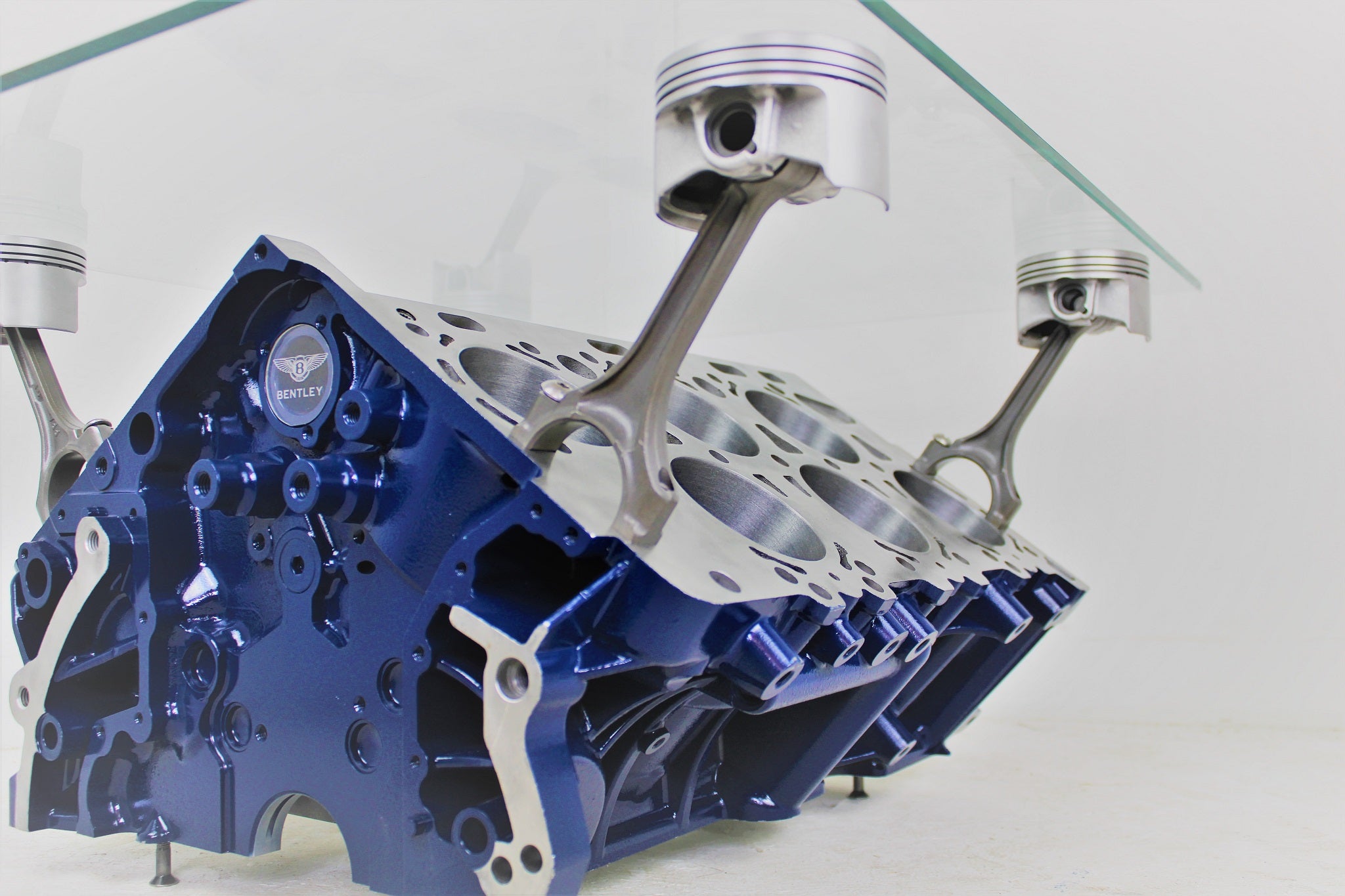 Bentley engine block coffee table with a rectangular glass top held up by car engine pistons. The table is painted dark blue, with the Bentley logo displayed on the front.