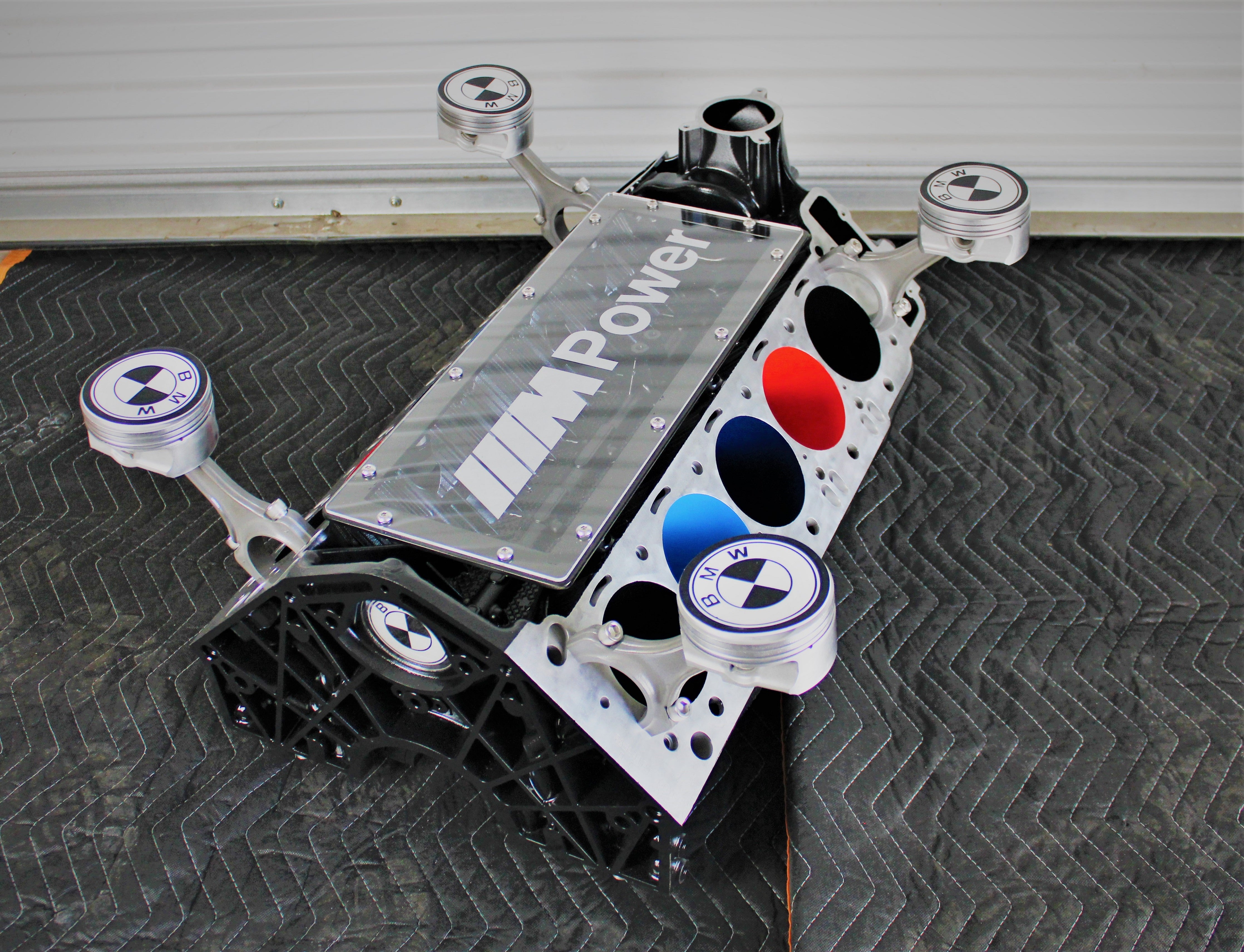 BMW M Series V10 engine block coffee table painted in the BMW M-Power color scheme without its glass top, the M-Power logo displayed across the piece, and the BMW logo etched into each of the four car pistons.