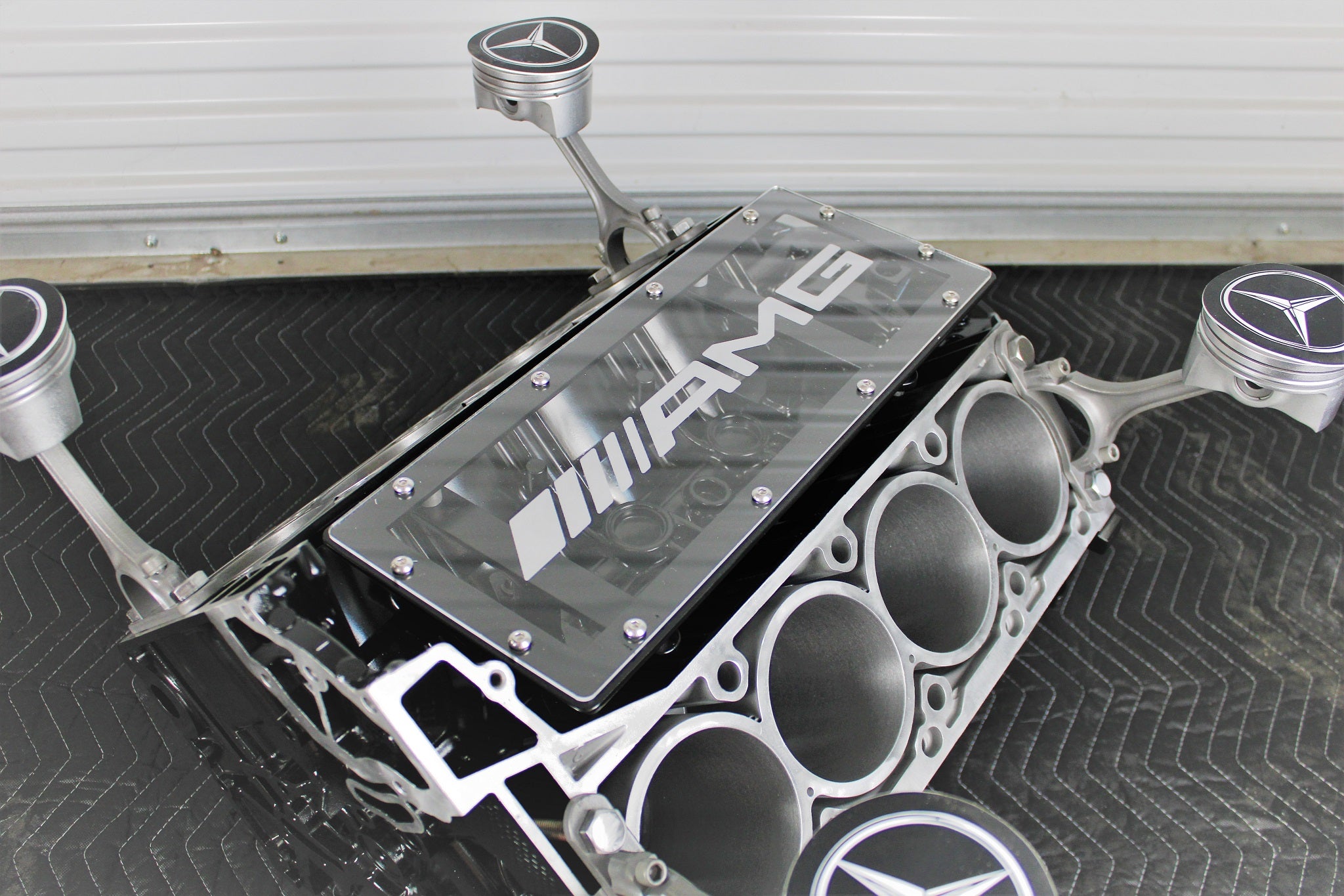 Engine block coffee table finished in black and silver, with the mercedes logo on each supporting piston. The AMG logo is displayed in the middle of the table without its glass top.