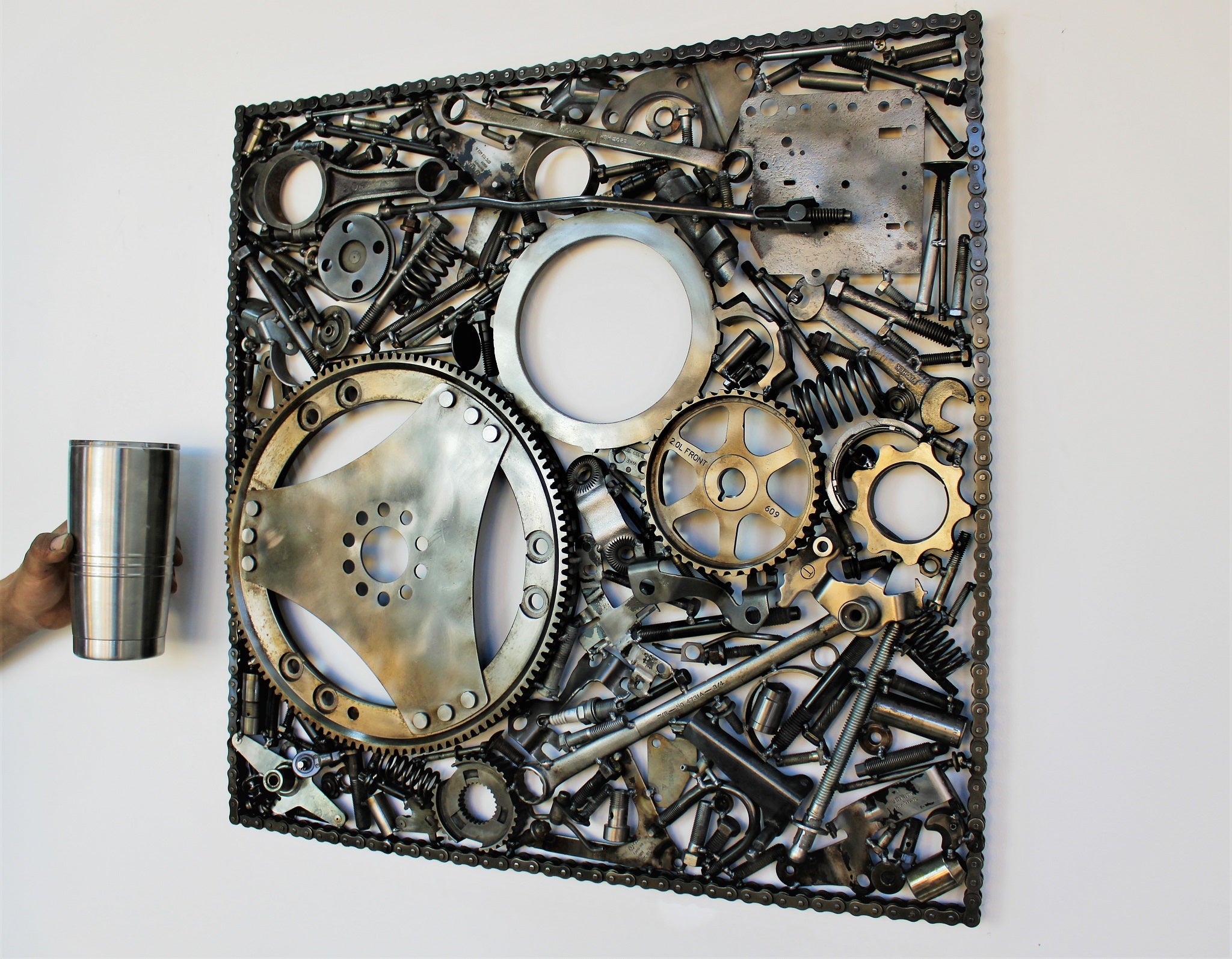 Automotive wall art made out of real car parts, 24 by 24 inch square wall hanging, next to a metal thermos for scale