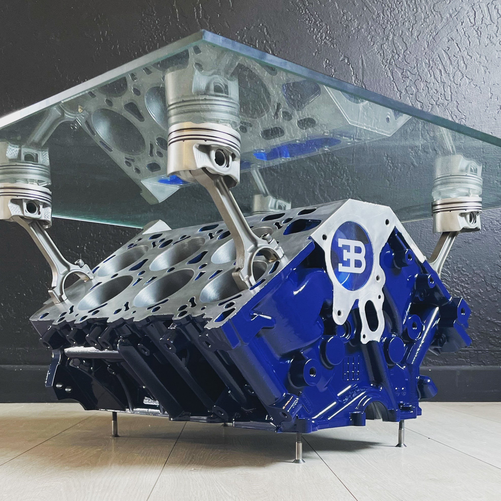Engine block coffee table painted blue with its square glass top being held up by car engine pistons.