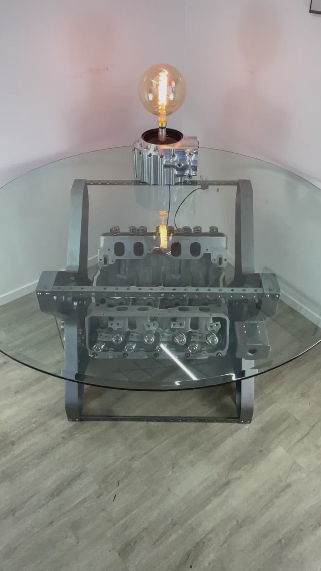 Video of an X-frame rotating engine dining table with a round glass top and car part lamp on top.