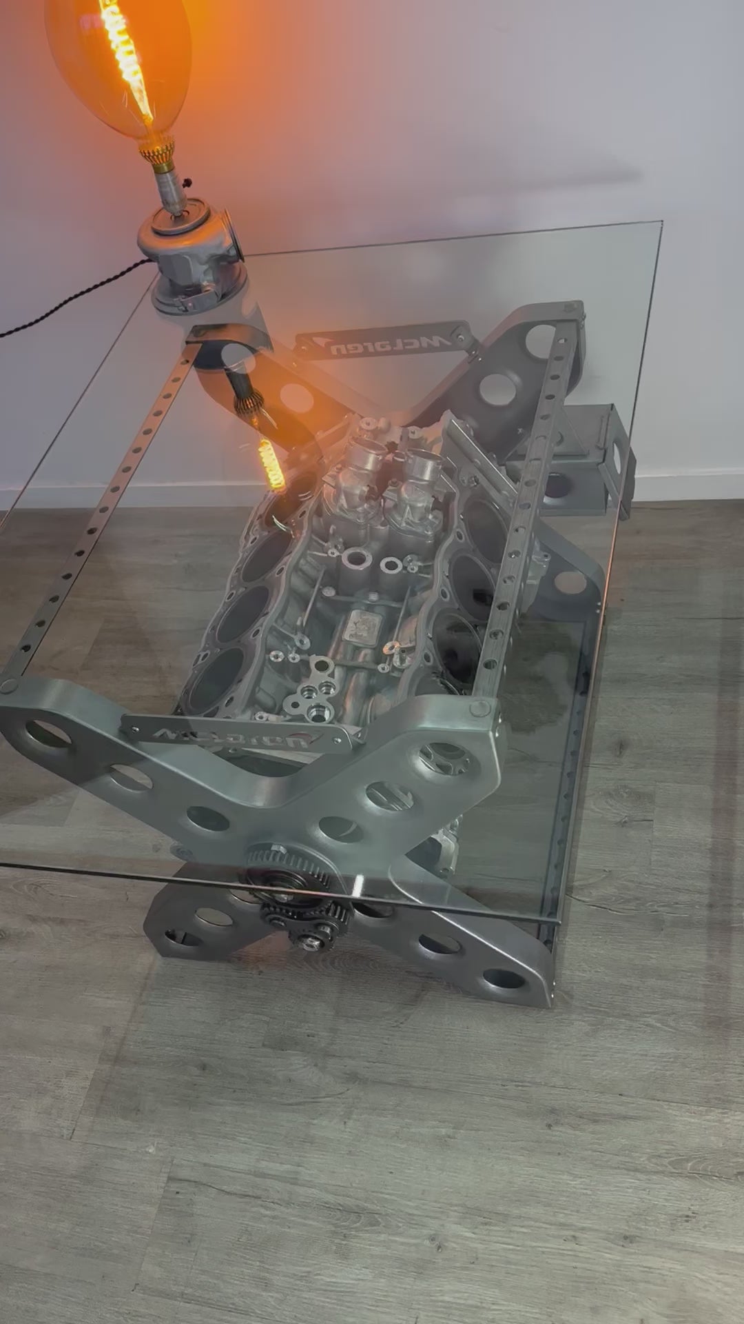 Video of a rotating engine X-frame coffee table, finished in silver with a rectangular glass top. The McLaren logo is displayed on both sides, and a car part lamp is lit on top of the table.