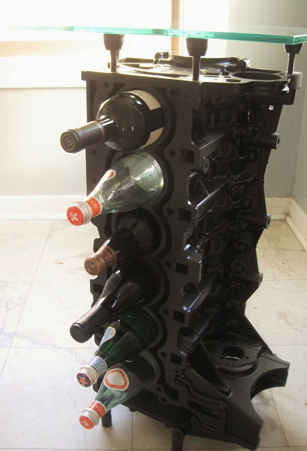 Engine block wine rack finished in black with a square glass top, bottles stored in its side.