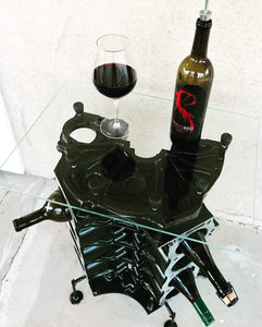 Birds-eye view of an engine block wine rack finished in black with a square glass top, wine bottles stored in its side and a wine bottle and glass on top.