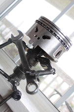 Load image into Gallery viewer, Piston man car part sculpture sitting on a windowsill and holding a wrench.
