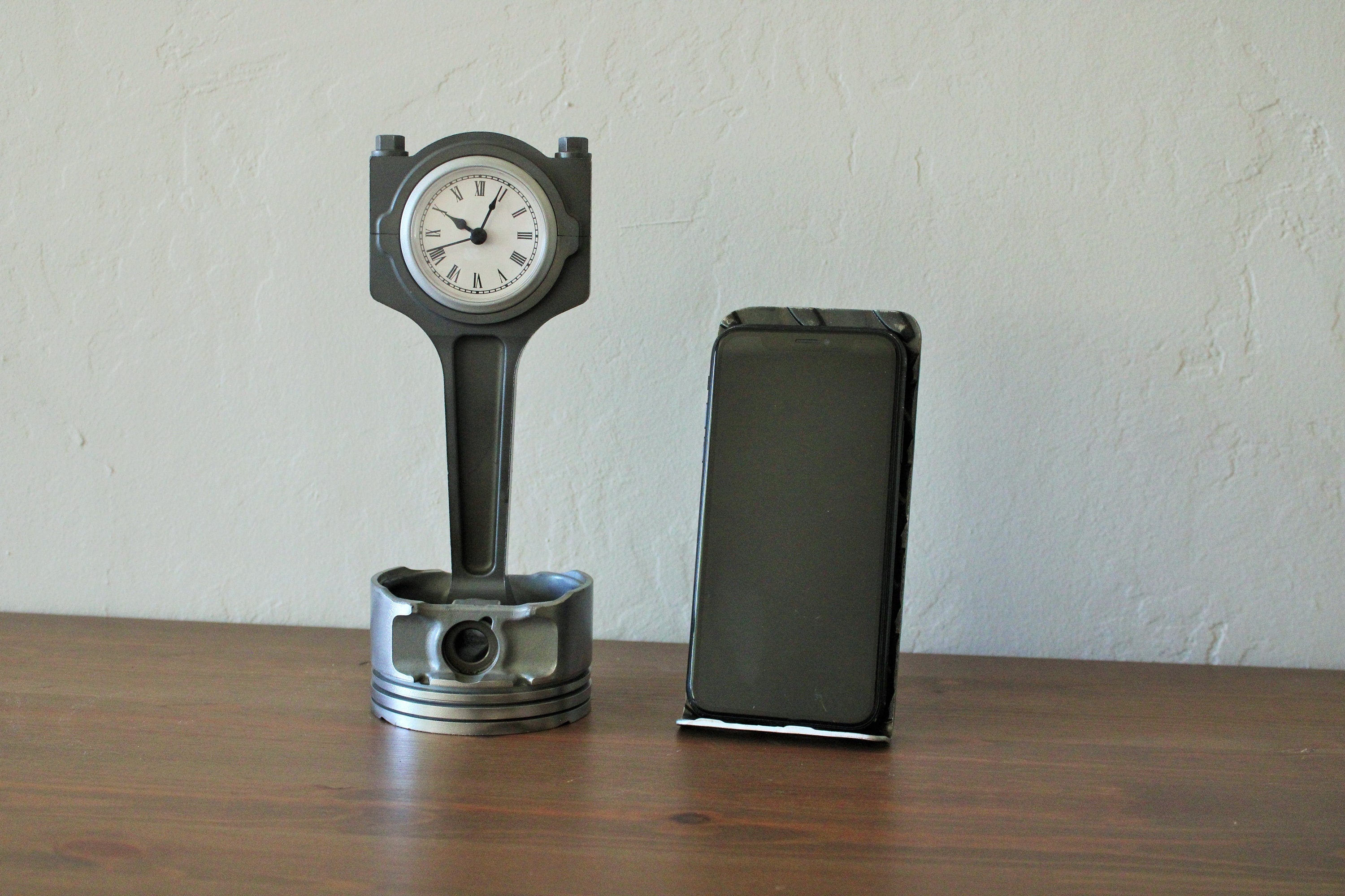 A piston clock made out of a Jaguar car's piston finished in gunmetal gray next to an iPhone on a stand.