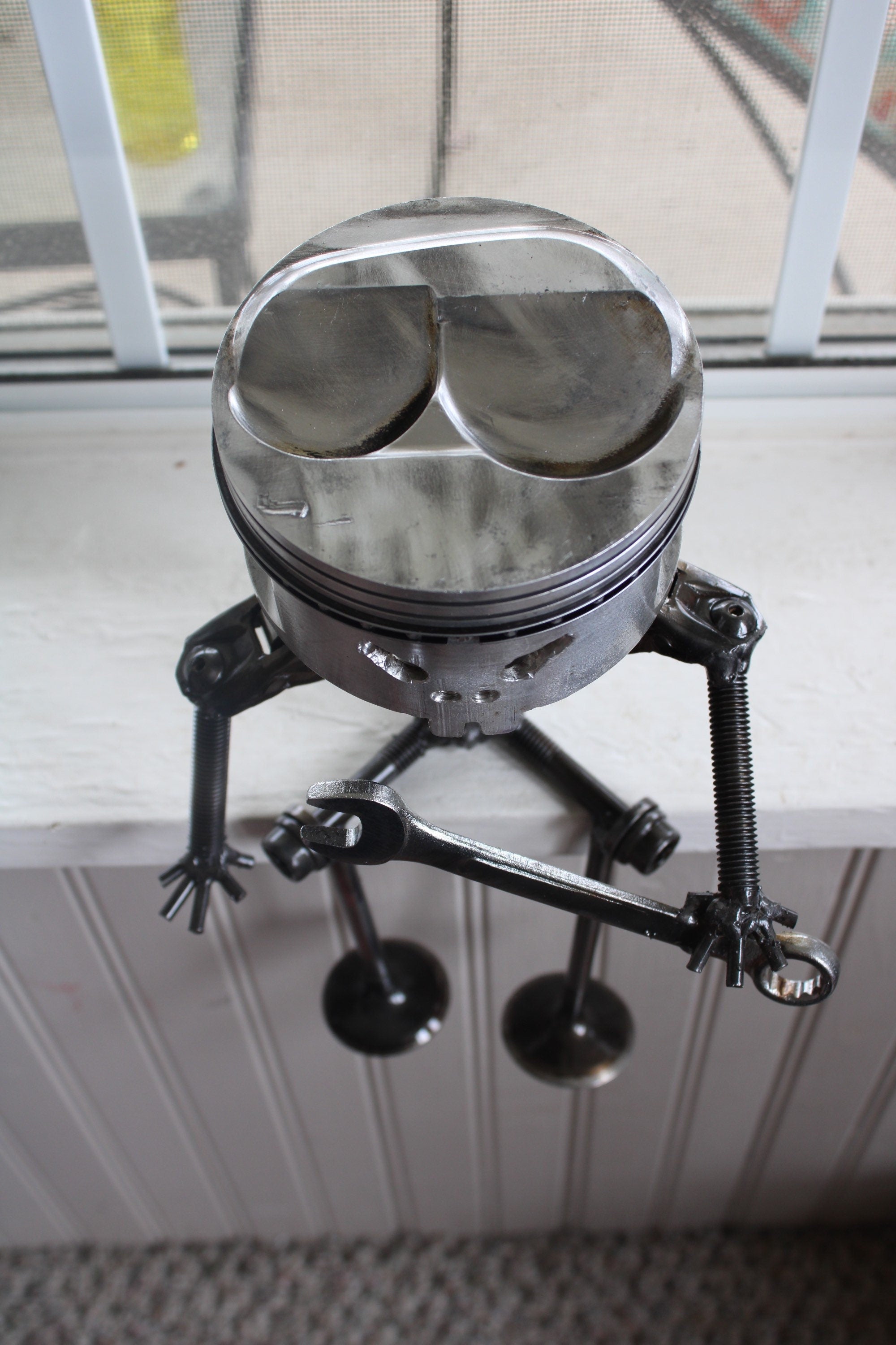 Birds-eye view of a piston man car part sculpture sitting on a windowsill and holding a wrench.